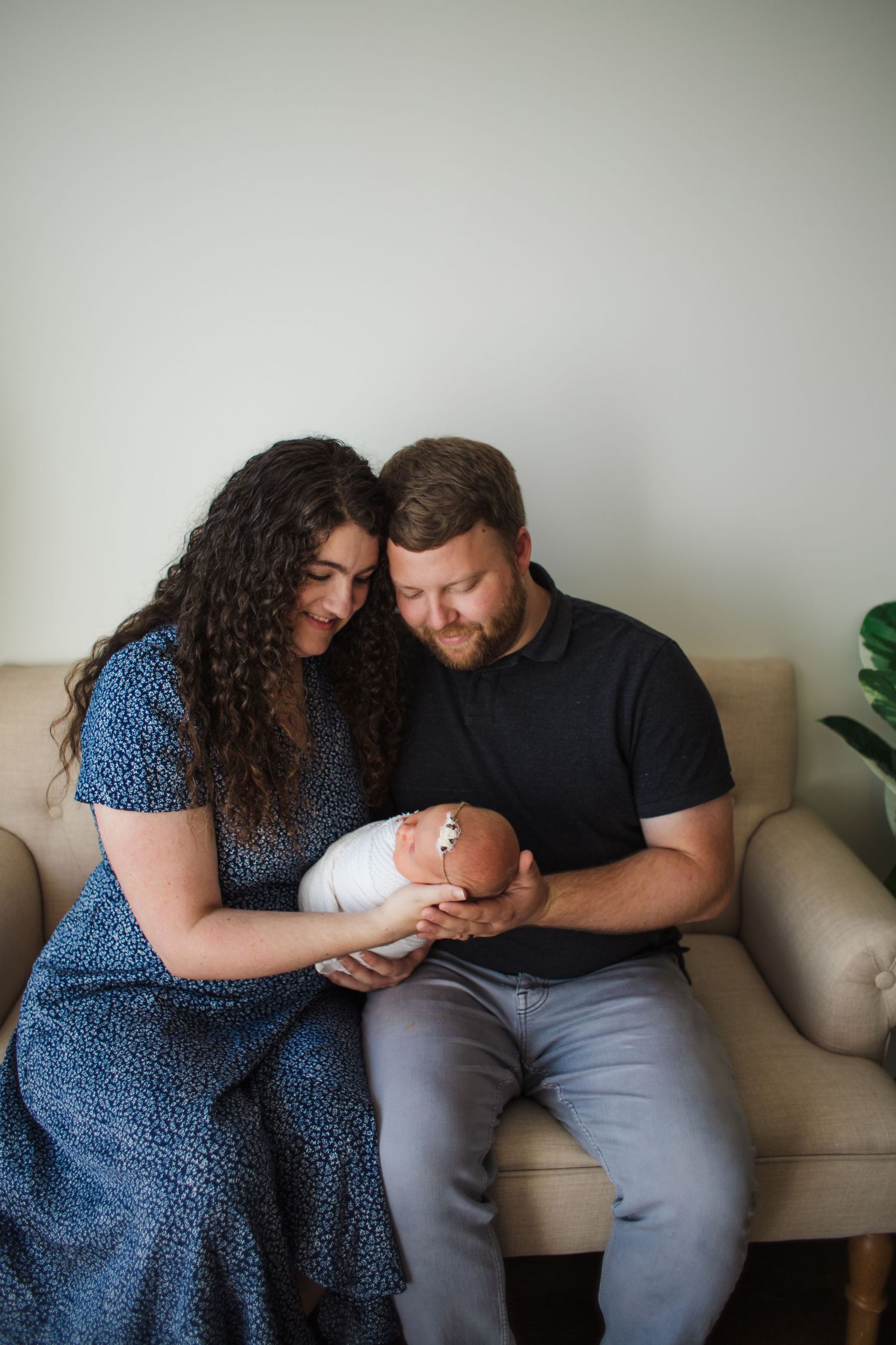 New parents sitting on a couch together lovingly gazing at their swaddled newborn baby.