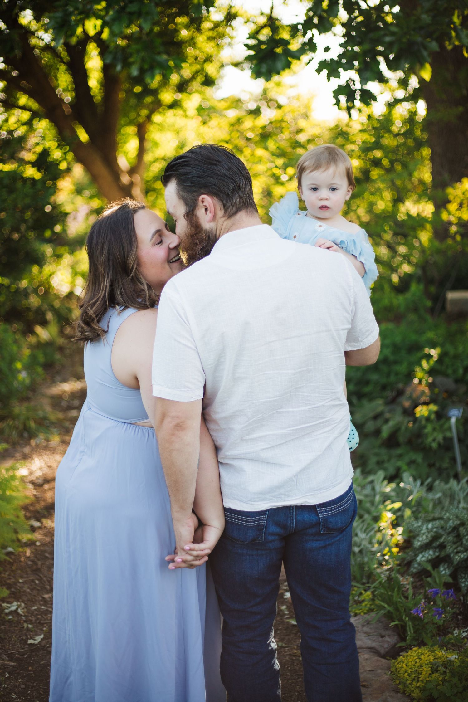 Couple sharing a tender moment while man holds their toddler in a lush green garden.