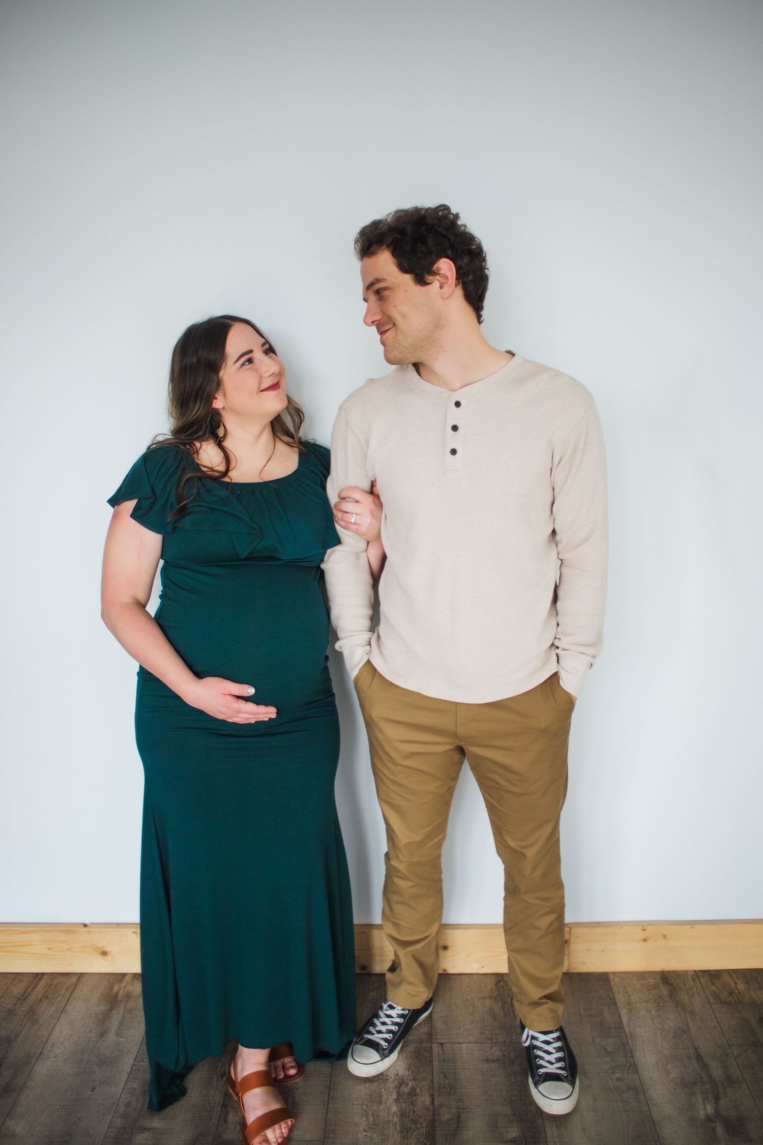 Pregnant woman in teal dress and partner smiling at each other against light blue wall.
