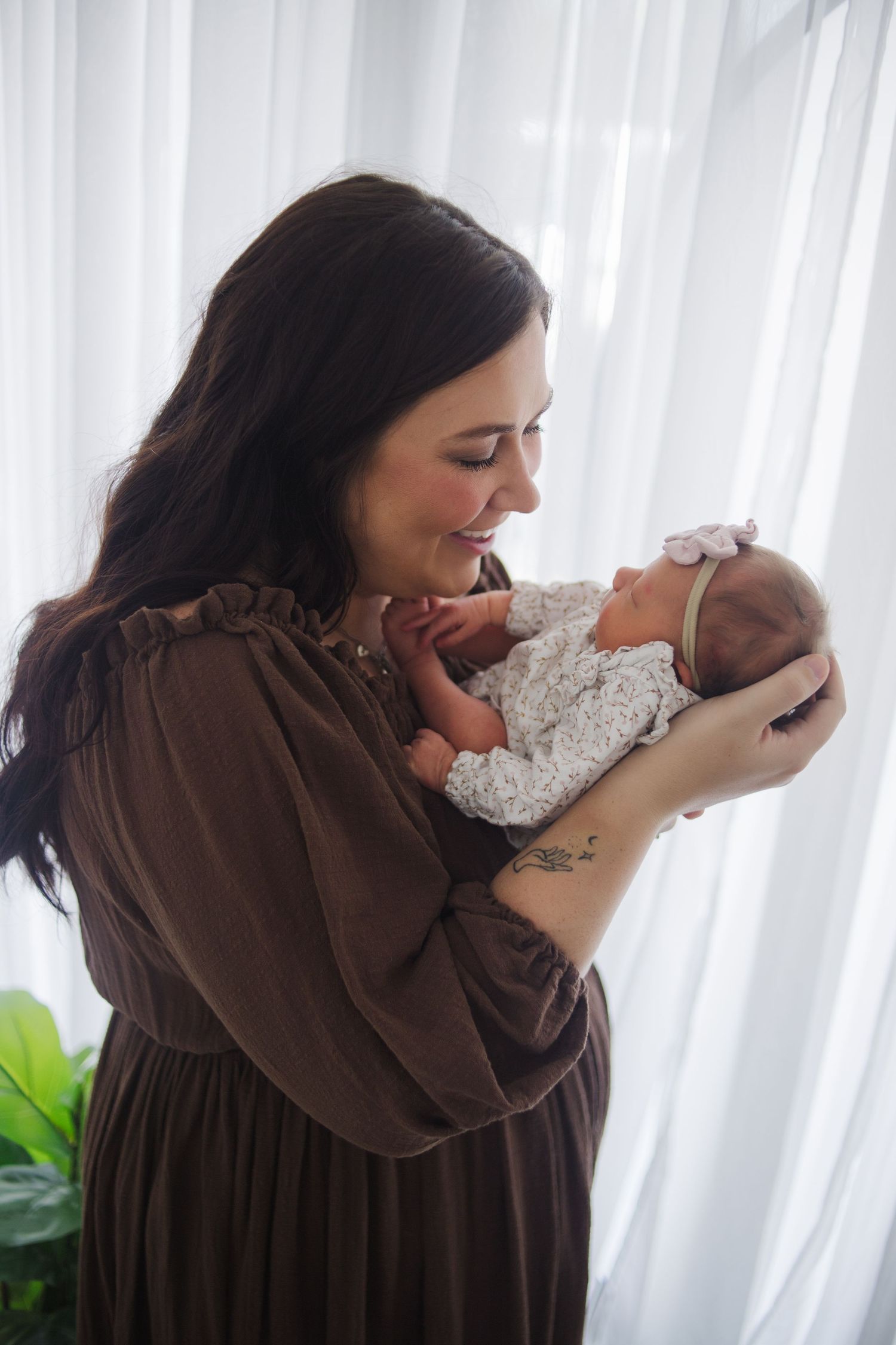 Dark-haired mother in brown dress smiling while gently holding her newborn baby by a window.