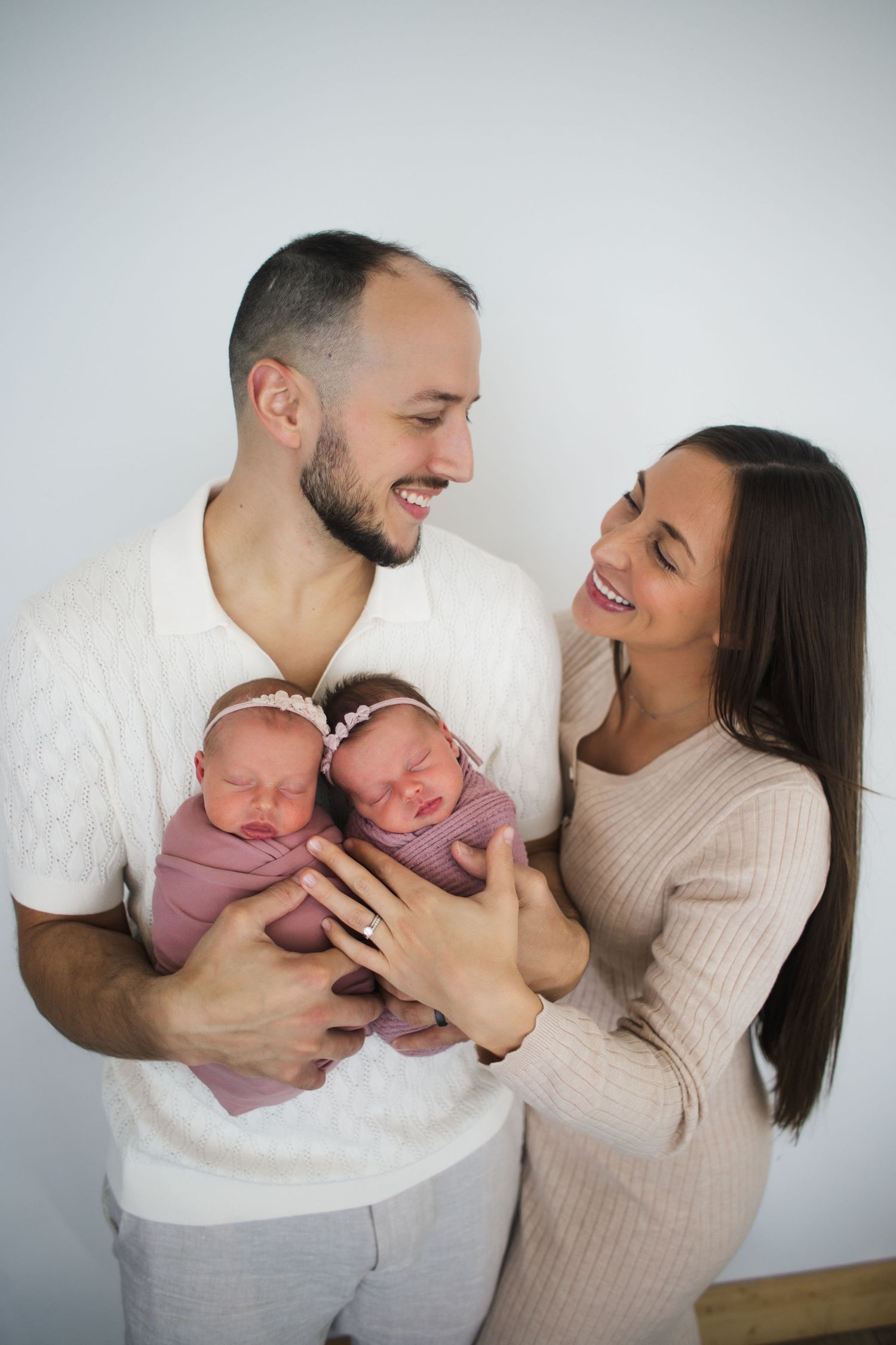 Smiling parents holding newborn twin girls wrapped in pink swaddles during a studio newborn photography session.