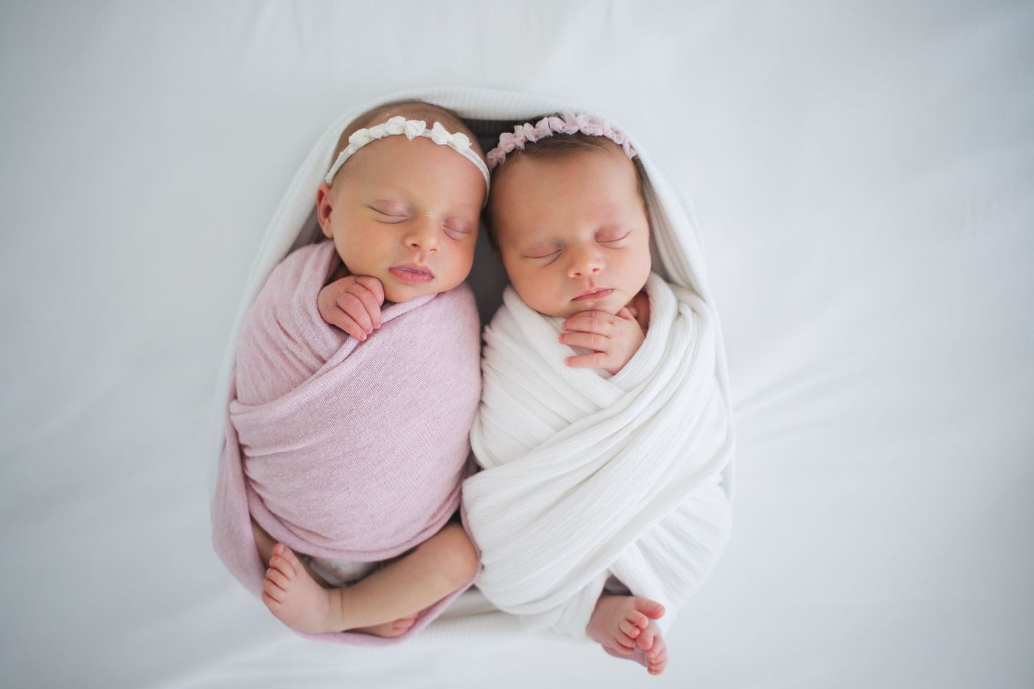 Twin newborn baby girls swaddled in pink and white wraps with floral headbands sleeping together.