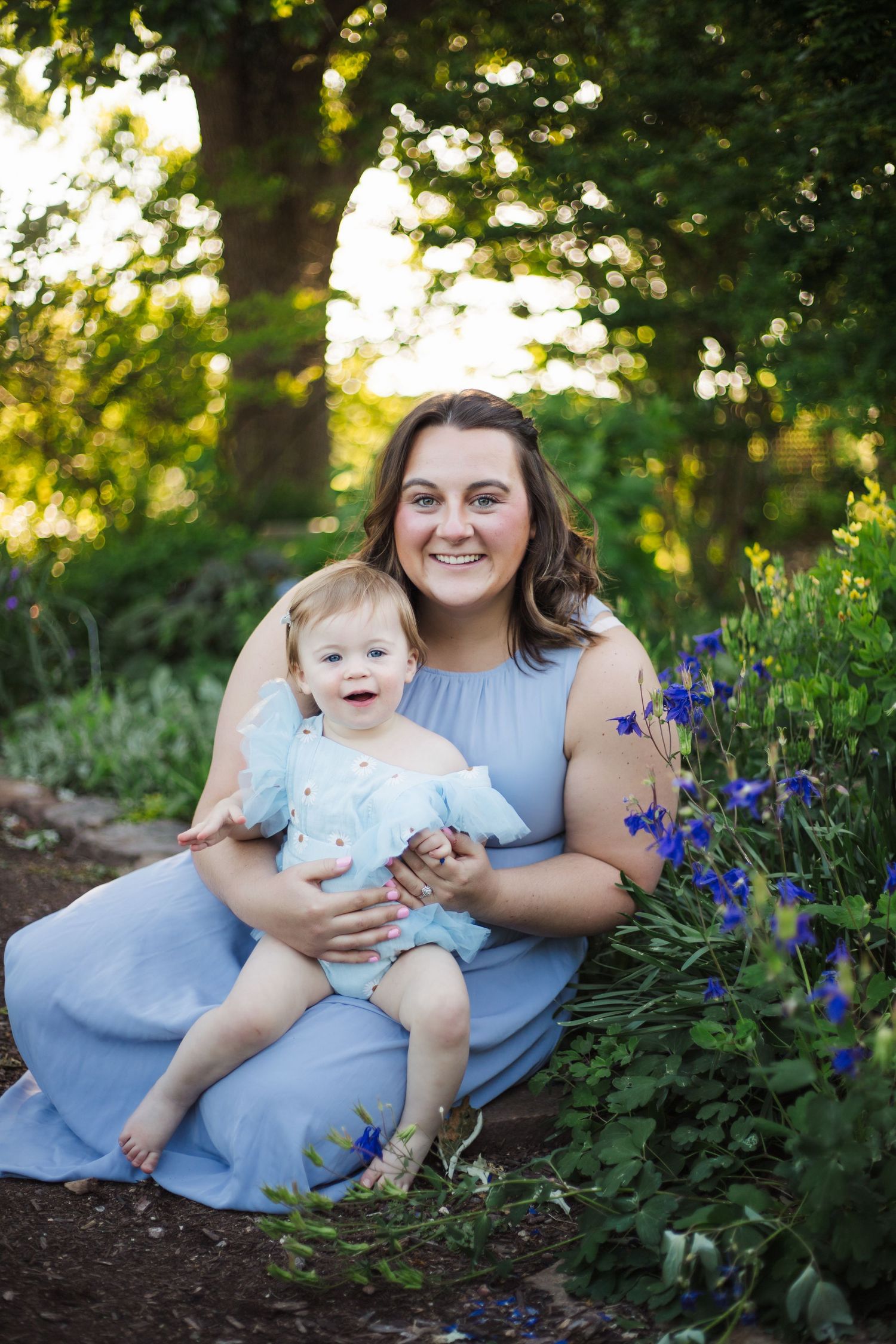 A smiling mother in a blue dress holds her happy baby girl among blue wildflowers in a garden.