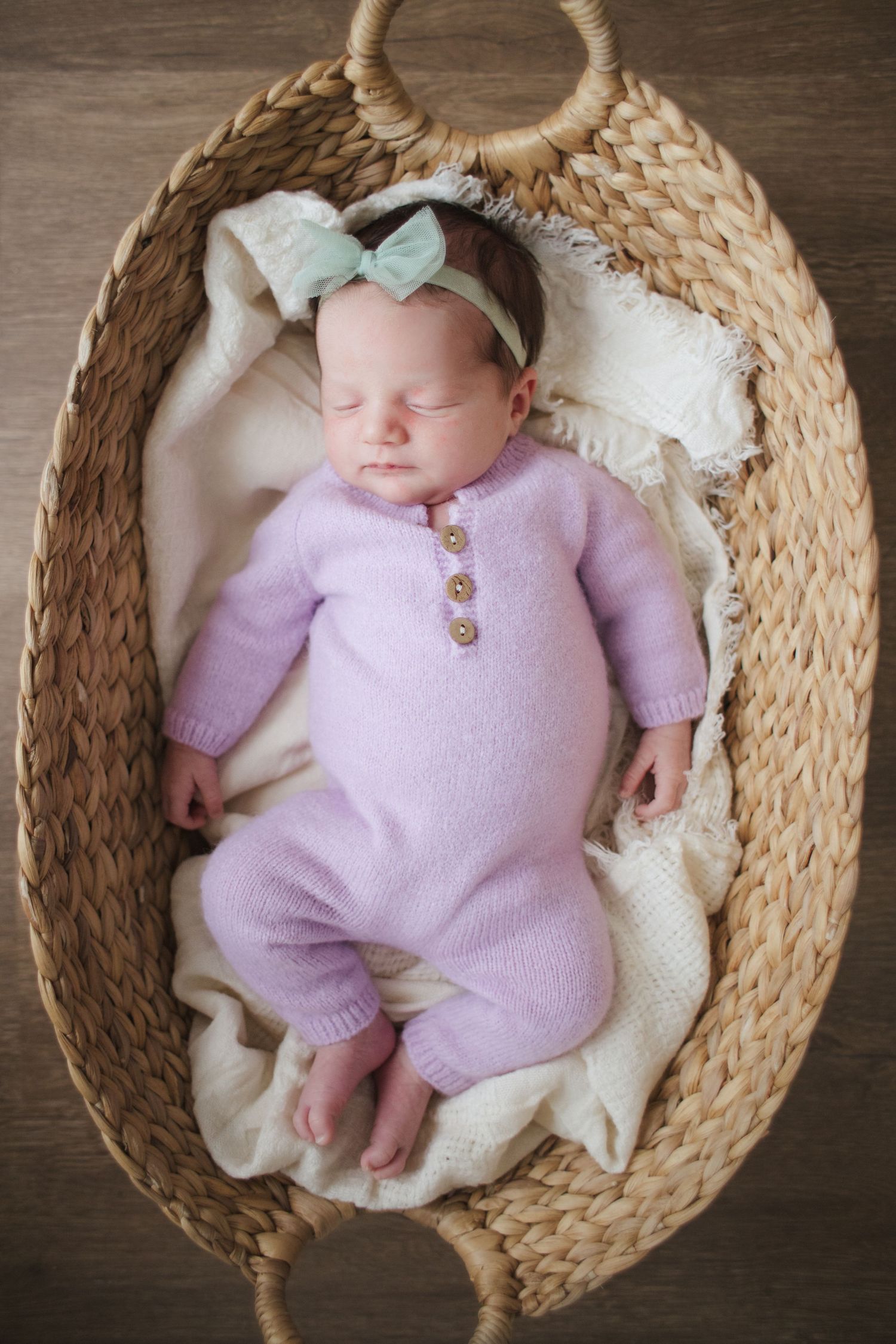 Sleeping newborn in lavender knit romper with mint bow headband lying in woven bassinet.