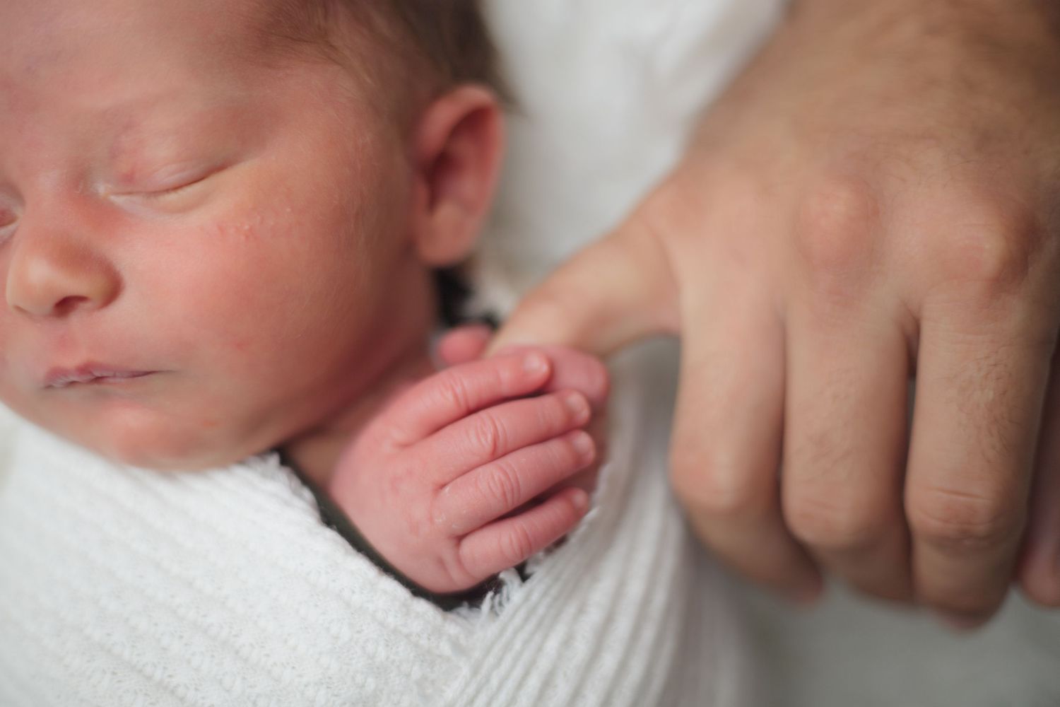 Closeup of a newborn baby grasping their fathers finger while swaddled in white.