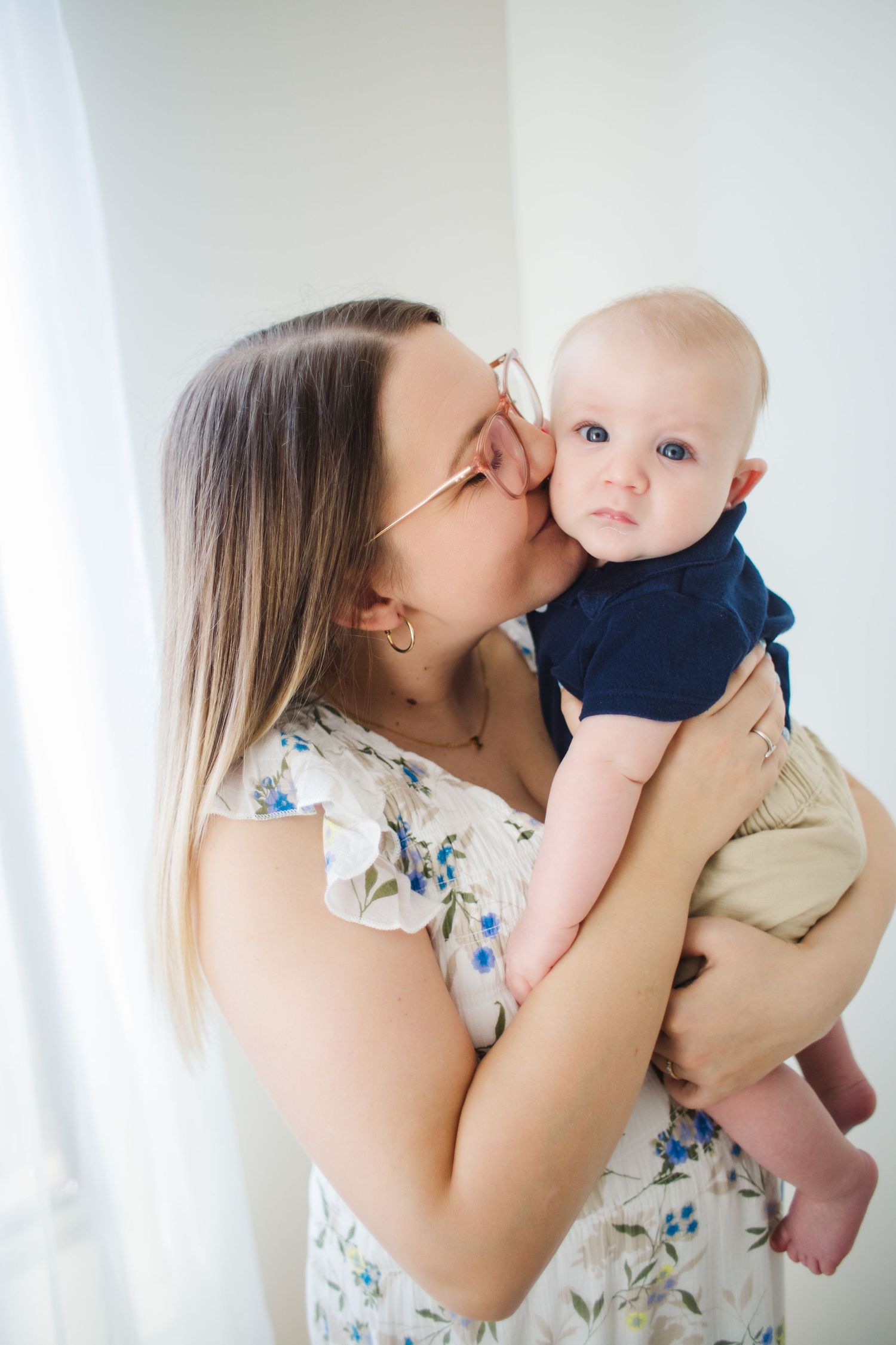 Young mother wearing glasses kissing her wide-eyed baby boy in bright white room.