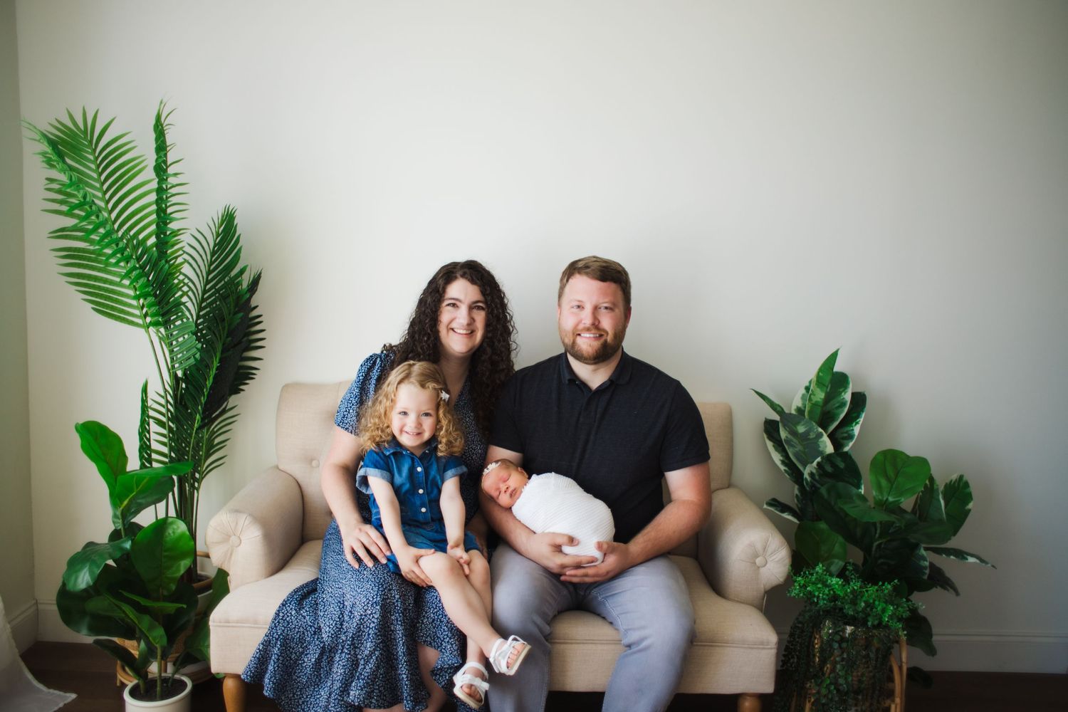 Family of four posing on a beige chair with newborn baby and toddler daughter surrounded by green plants.