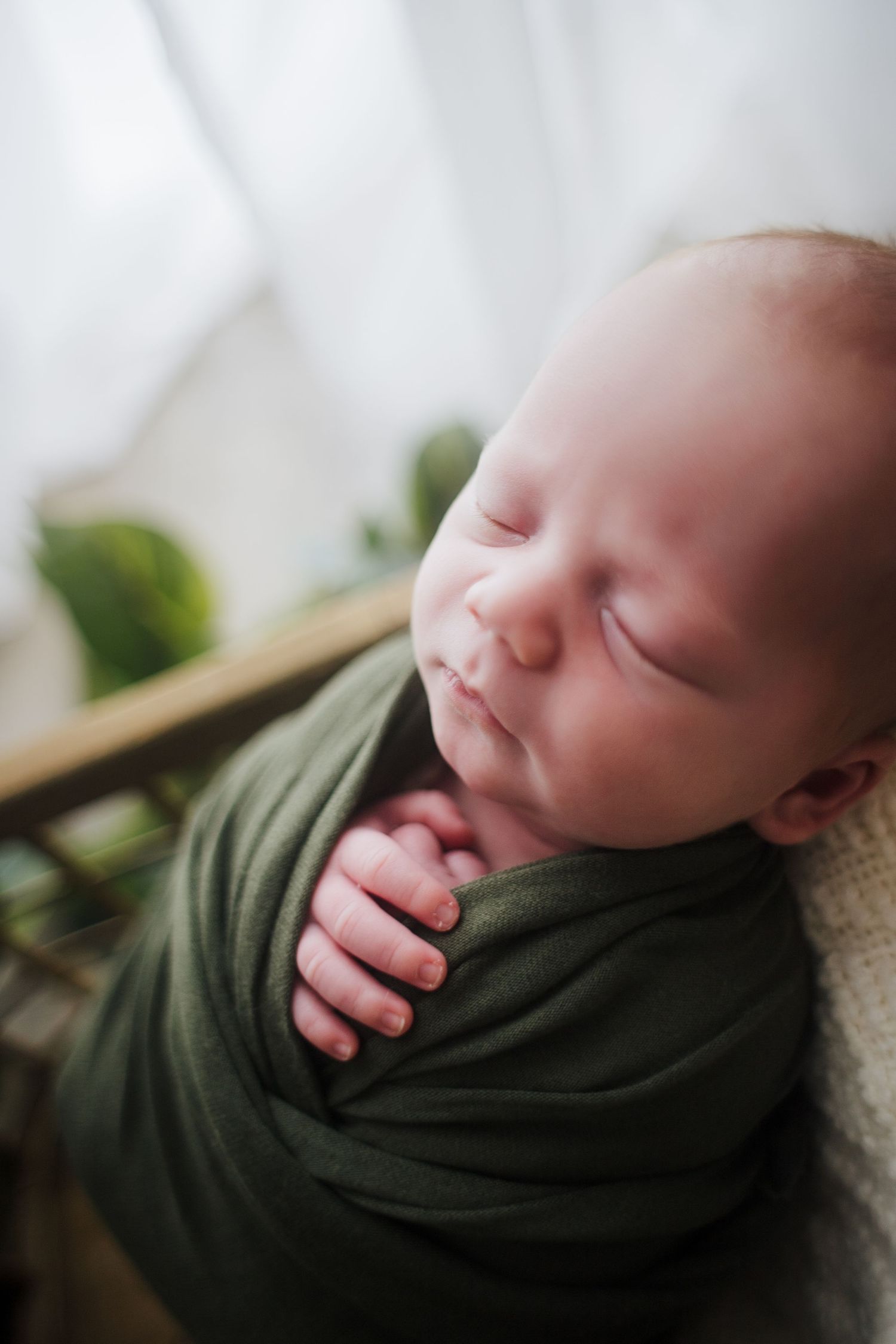 Peaceful sleeping newborn baby swaddled in olive green wrap with tiny hand peeking out.