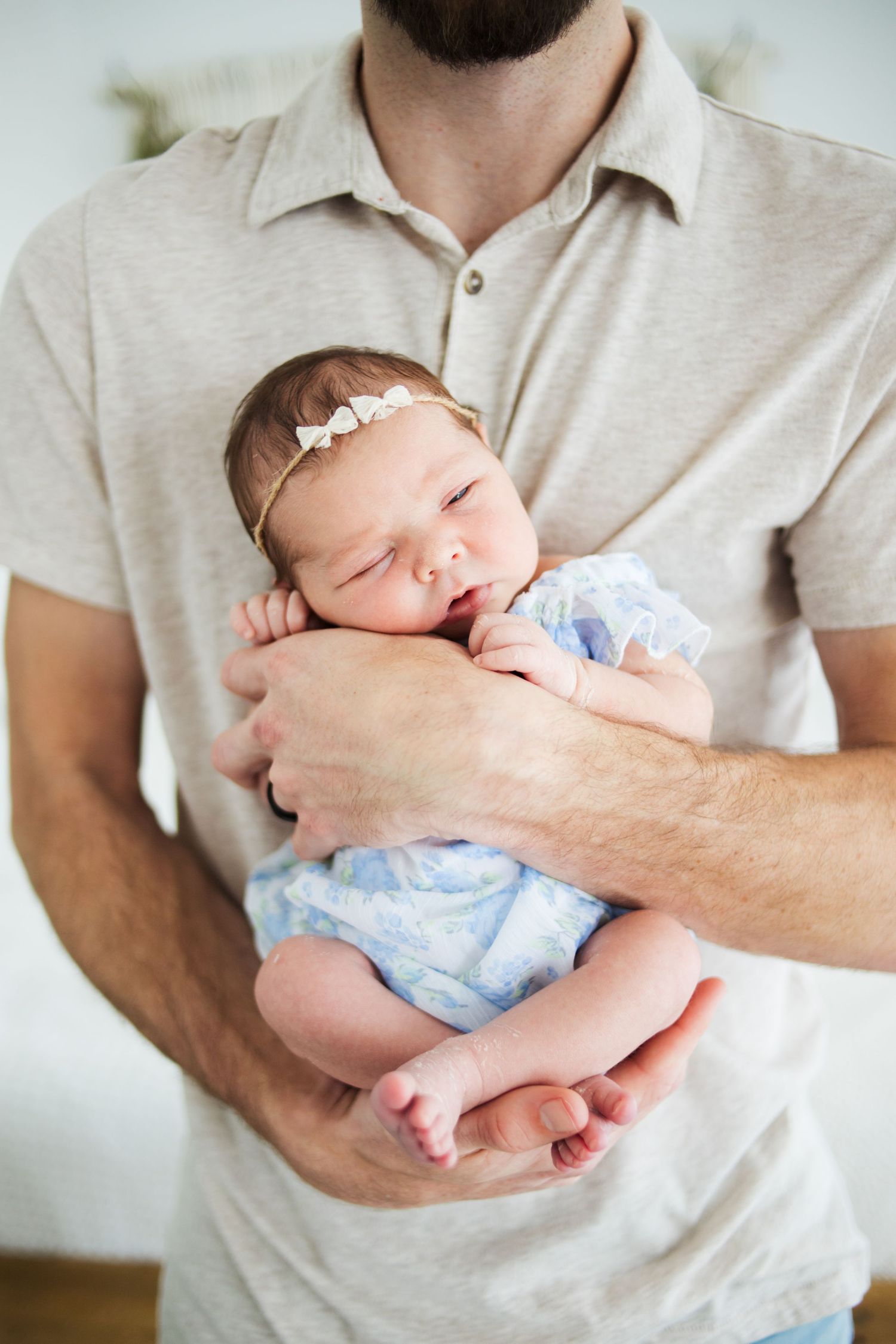 Father in beige polo shirt cradling sleeping newborn baby girl in blue outfit with headband.