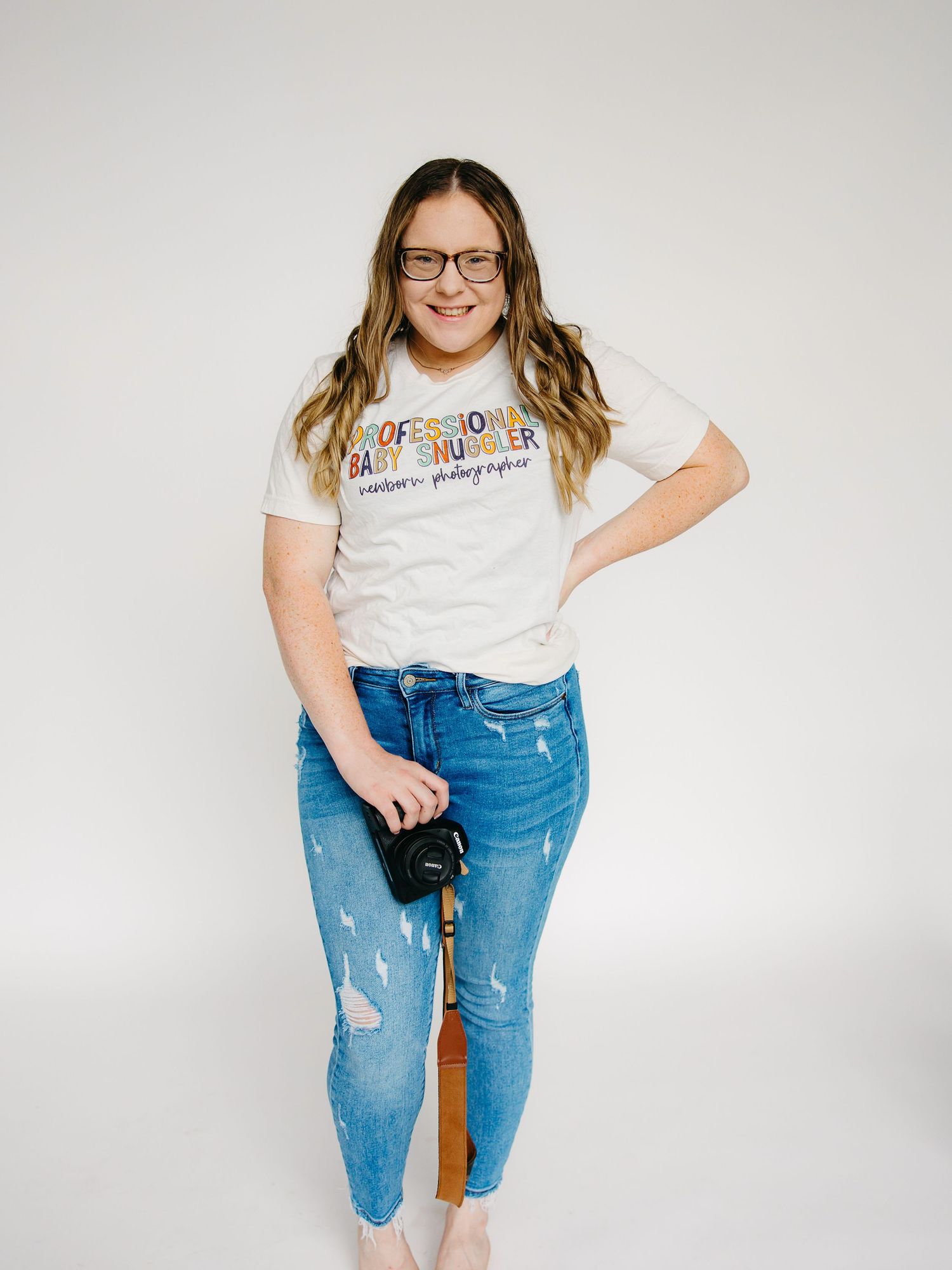 Female newborn photographer smiling in white professional baby snuggler t-shirt holding camera, wearing glasses.