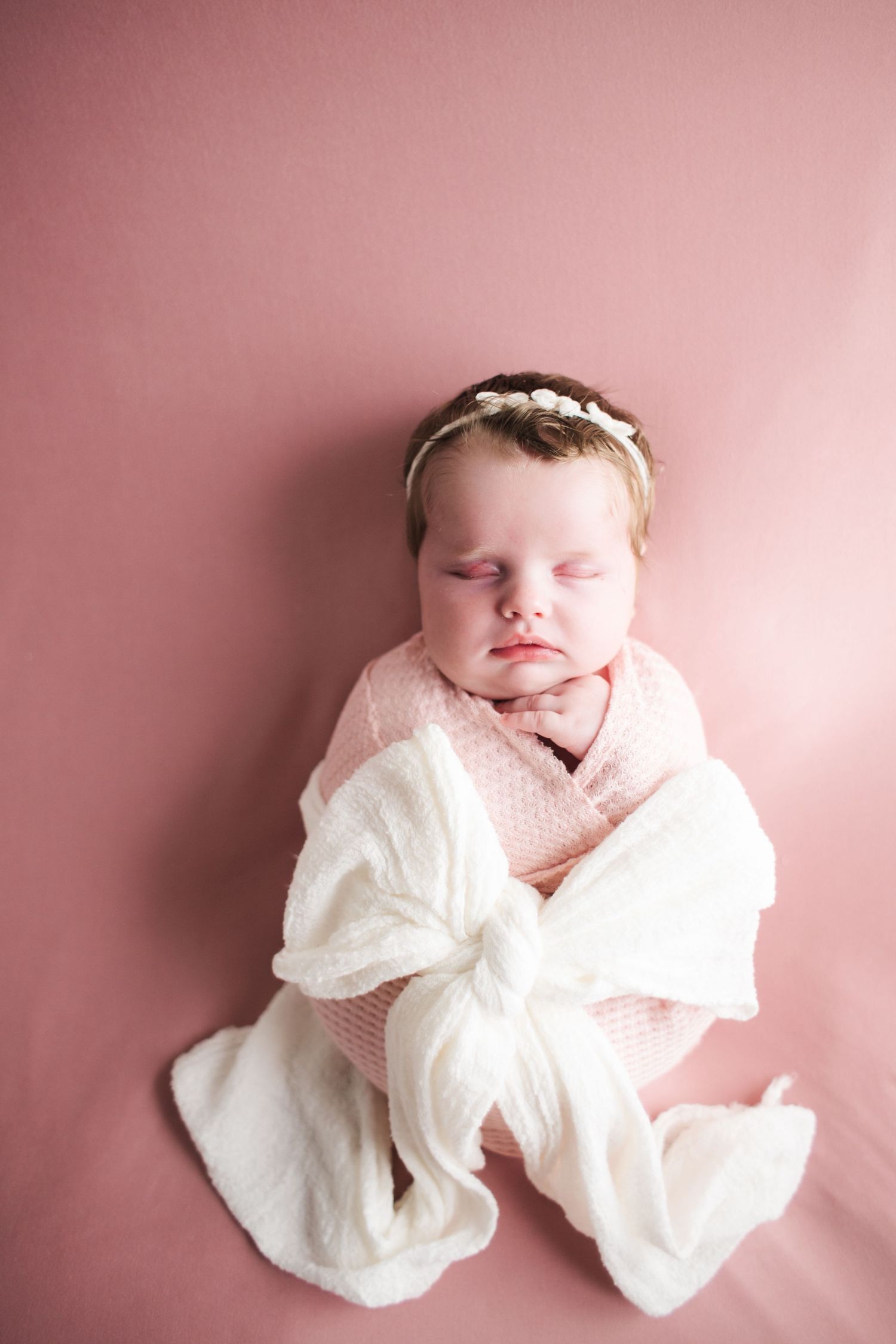 Sleeping newborn baby swaddled in pink wrap with white bow and floral headband on pink backdrop.