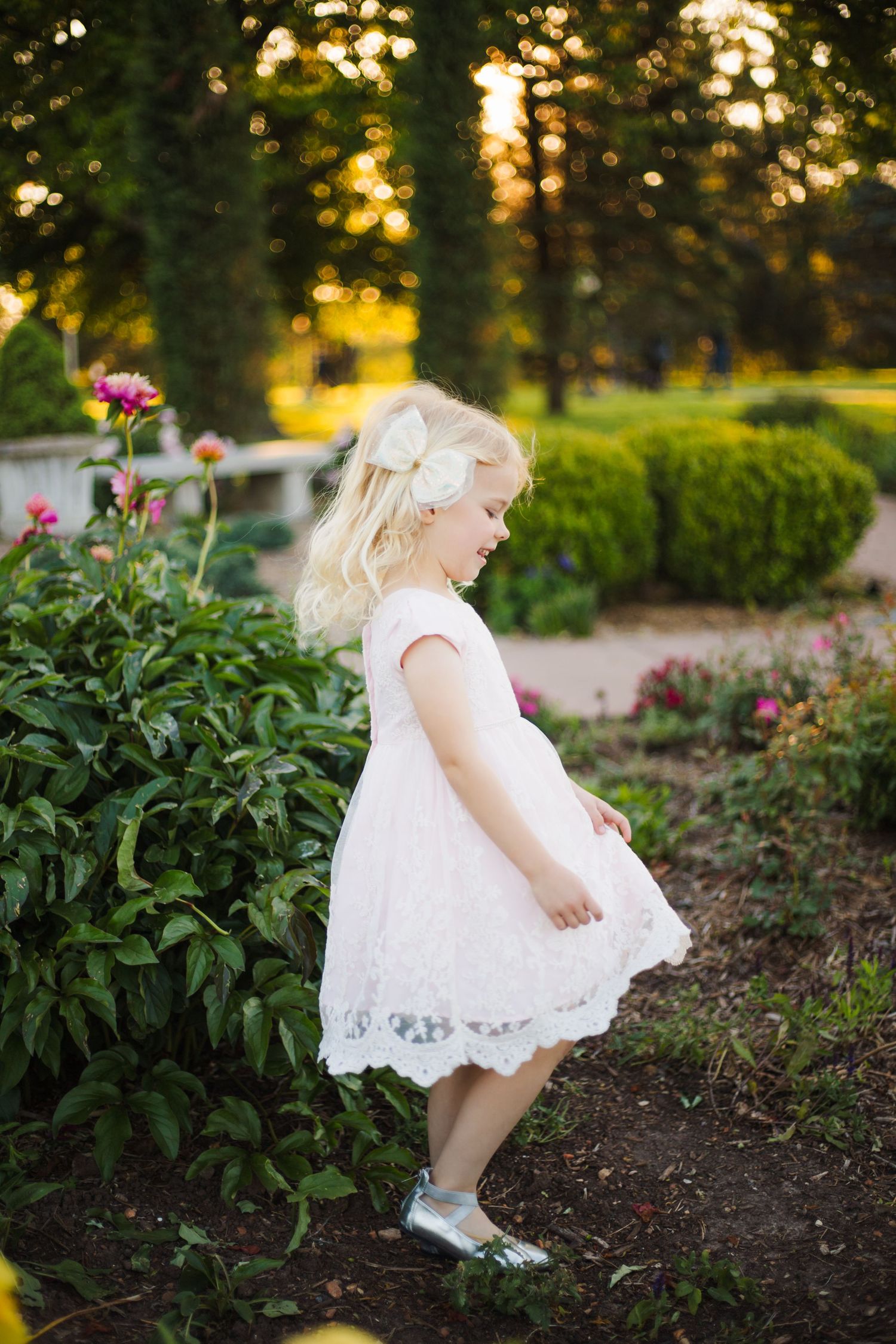 A blonde toddler girl in a white lace dress explores a lush garden at golden hour sunset.