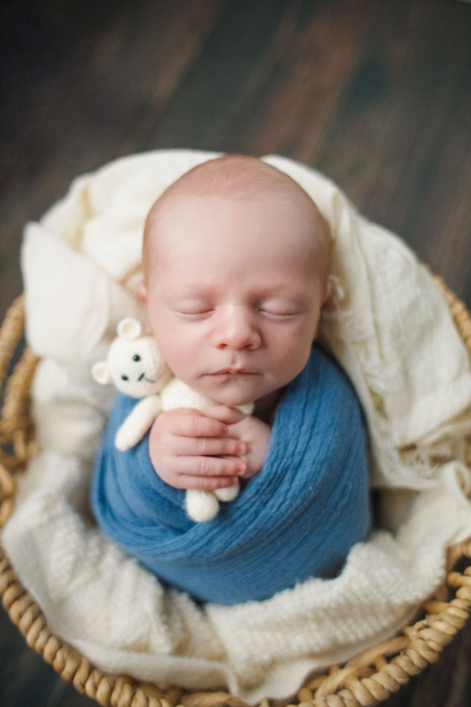 Sleeping newborn baby swaddled in blue wrap holding tiny teddy bear in wicker basket.