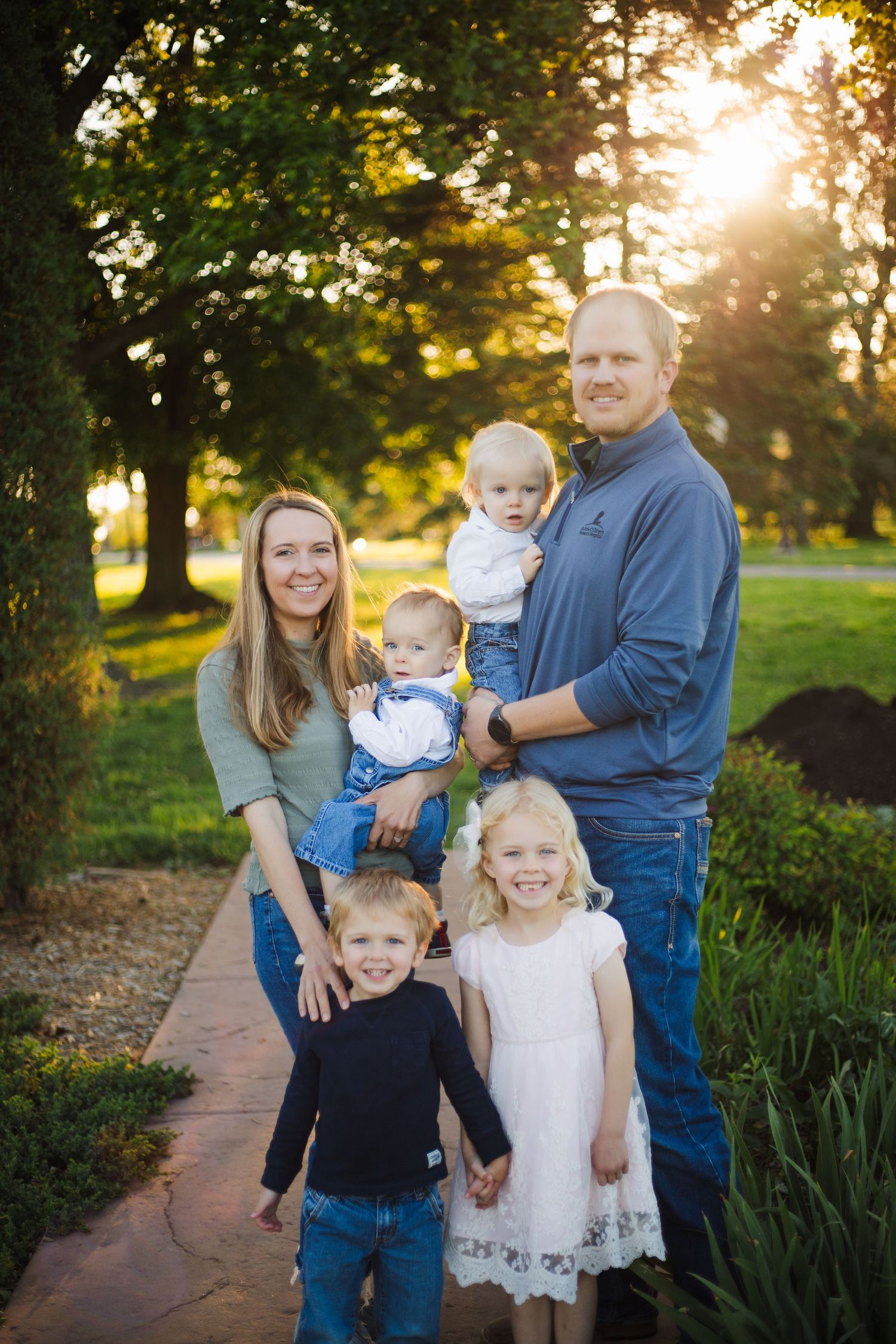 Family of six posing on a sunlit park path at sunset, parents holding two young children.