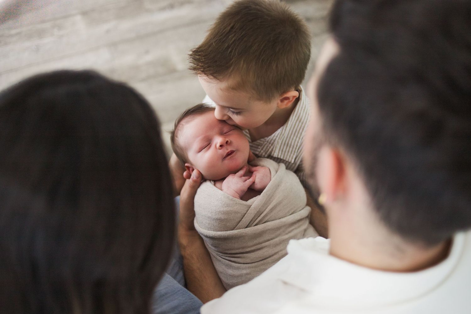 Proud big brother gently kisses sleeping newborn sibling held by parents.