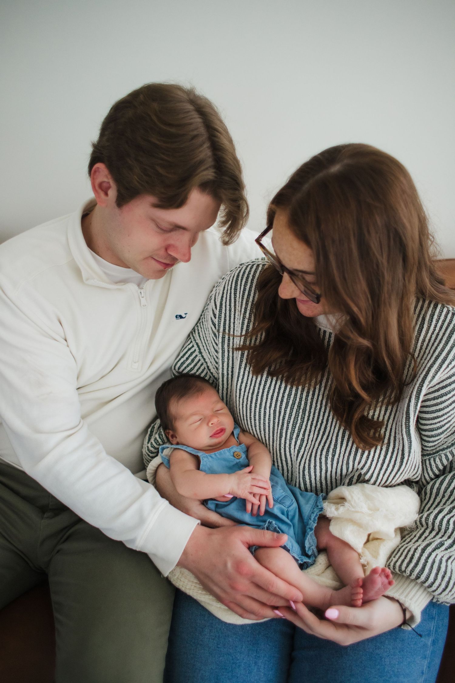 New parents in casual attire lovingly gazing at their sleeping newborn dressed in blue denim overalls.
