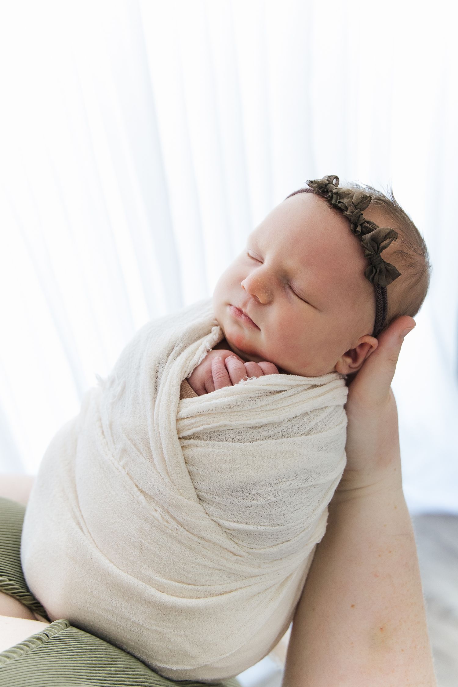 Peaceful sleeping newborn baby girl with floral headband swaddled in cream wrap cradled in parent's hands.