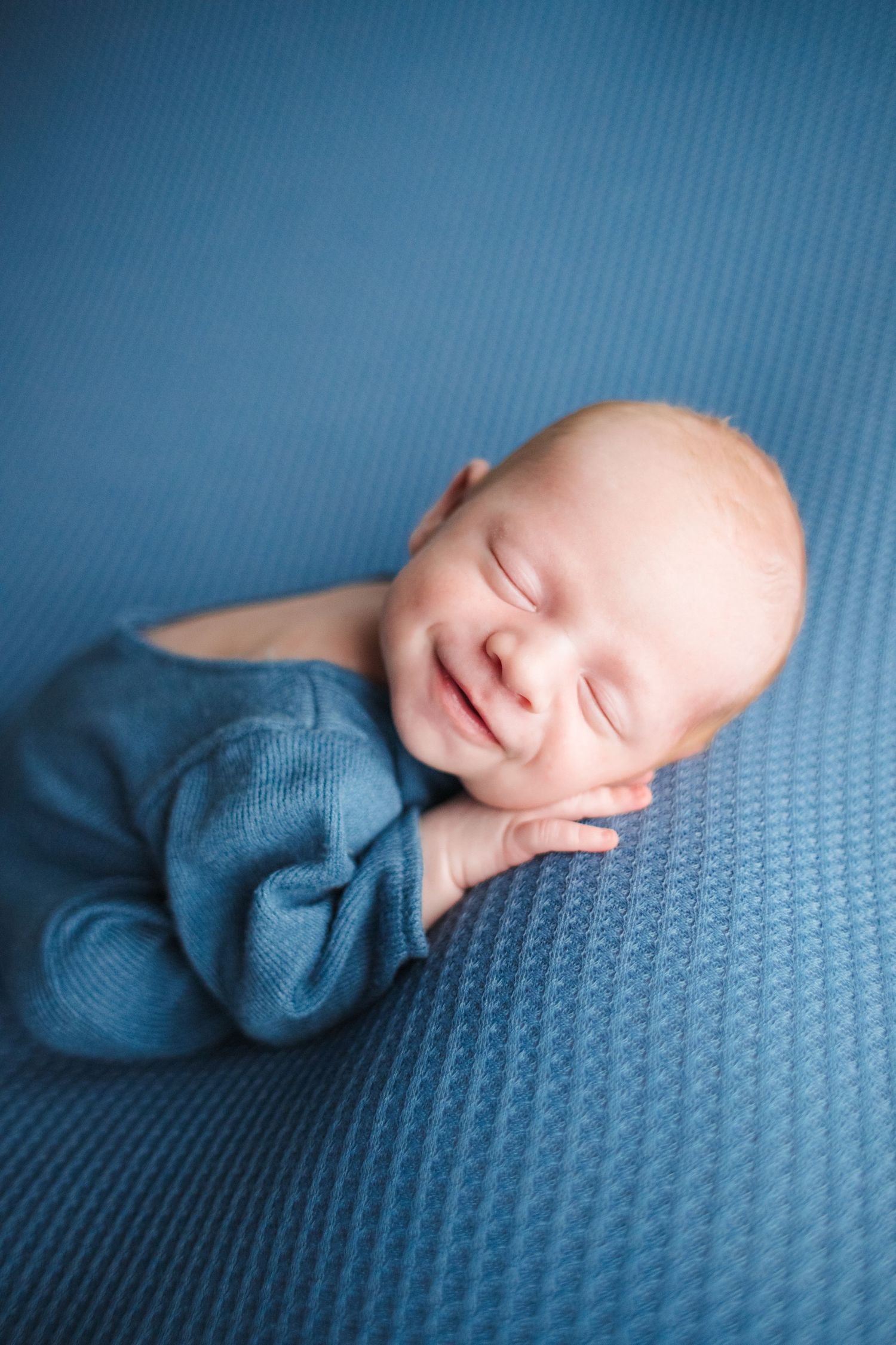 Smiling newborn baby boy in blue knit outfit lying on matching blue textured blanket during photo session.