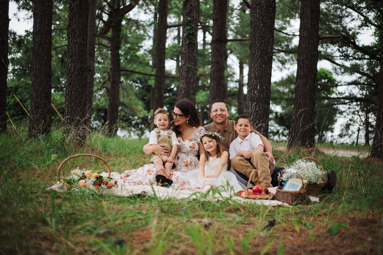 Happy family of six enjoying a woodland picnic surrounded by tall pine trees with a flower basket nearby.