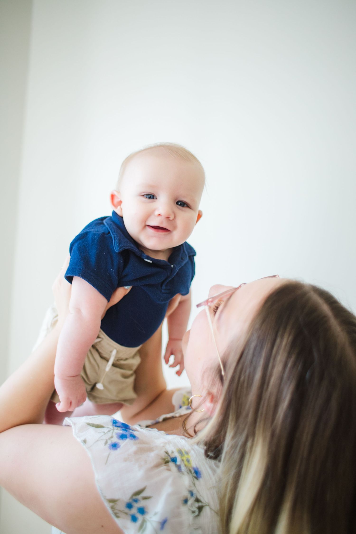 A joyful baby in a blue shirt smiles while being lifted by a mother indoors.
