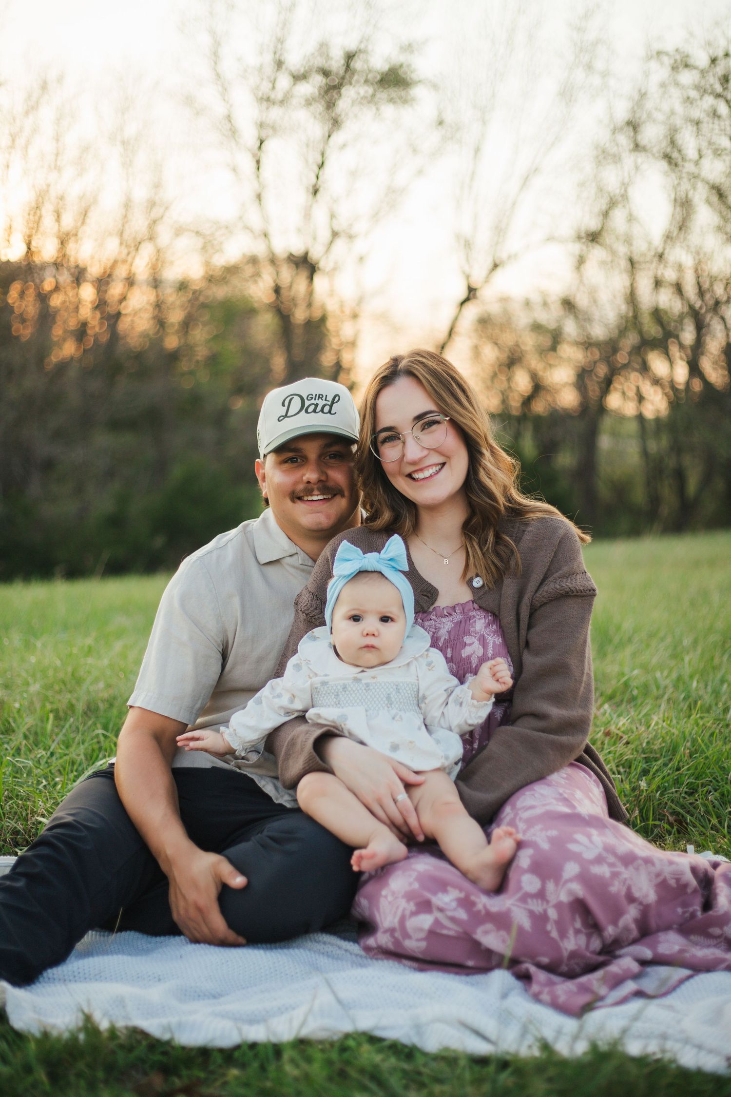 Smiling young family sitting on a blanket in a grassy field at sunset with their baby girl.