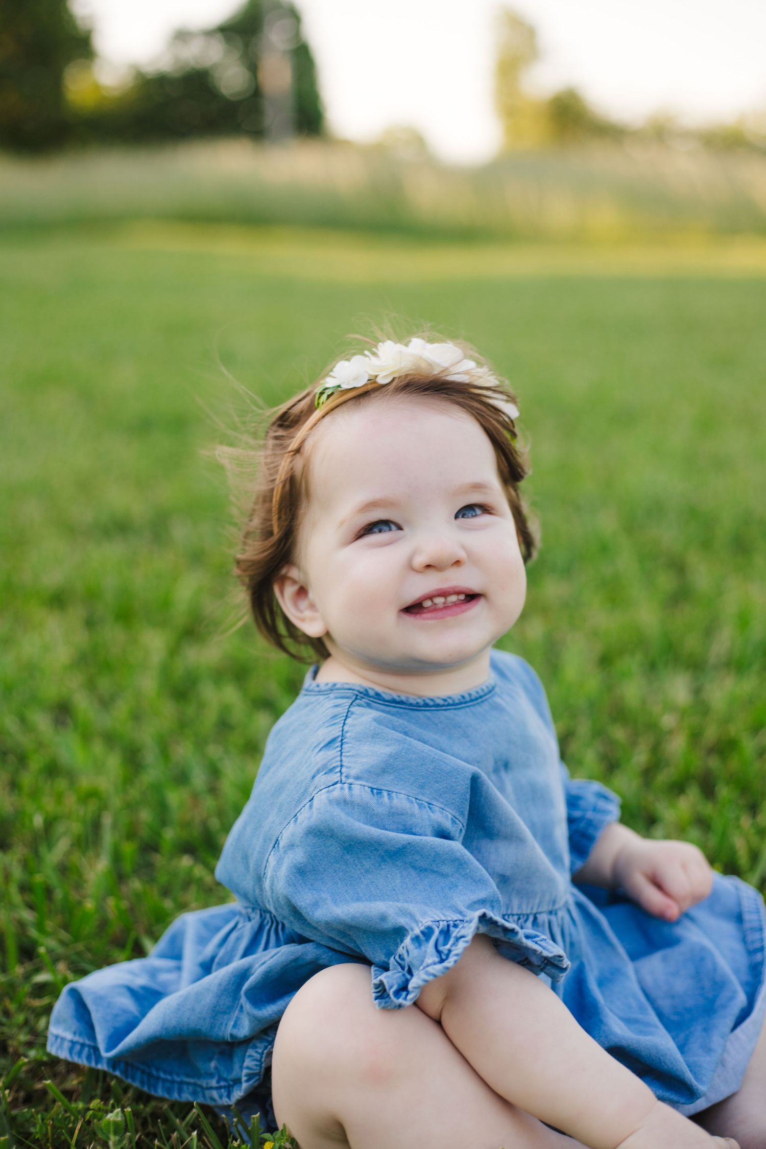 Happy toddler girl in blue denim dress with flower headband sitting on green grass smiling.