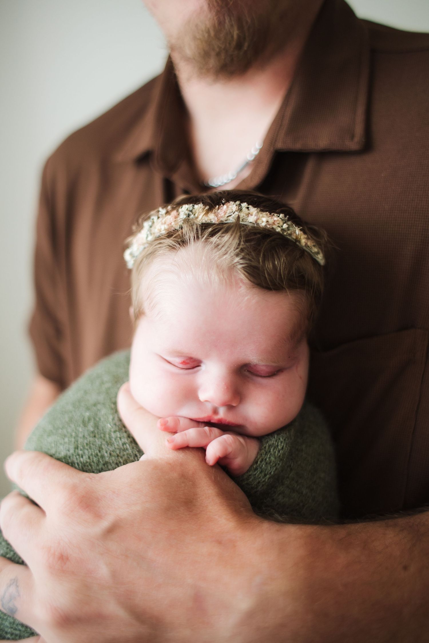 Father in brown shirt gently cradling sleeping newborn baby wrapped in green with floral headband.