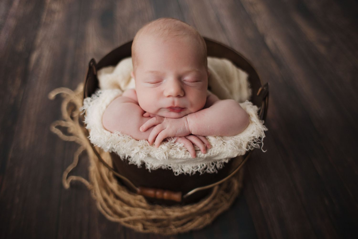 Sleeping newborn baby with hands under chin posed in a rustic wooden bucket on dark wood floor.