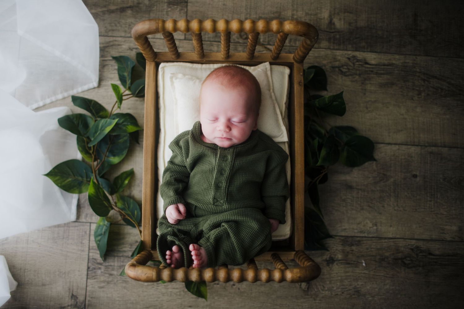 Sleeping newborn baby in green romper lying in small wooden beaded crib with greenery decor.