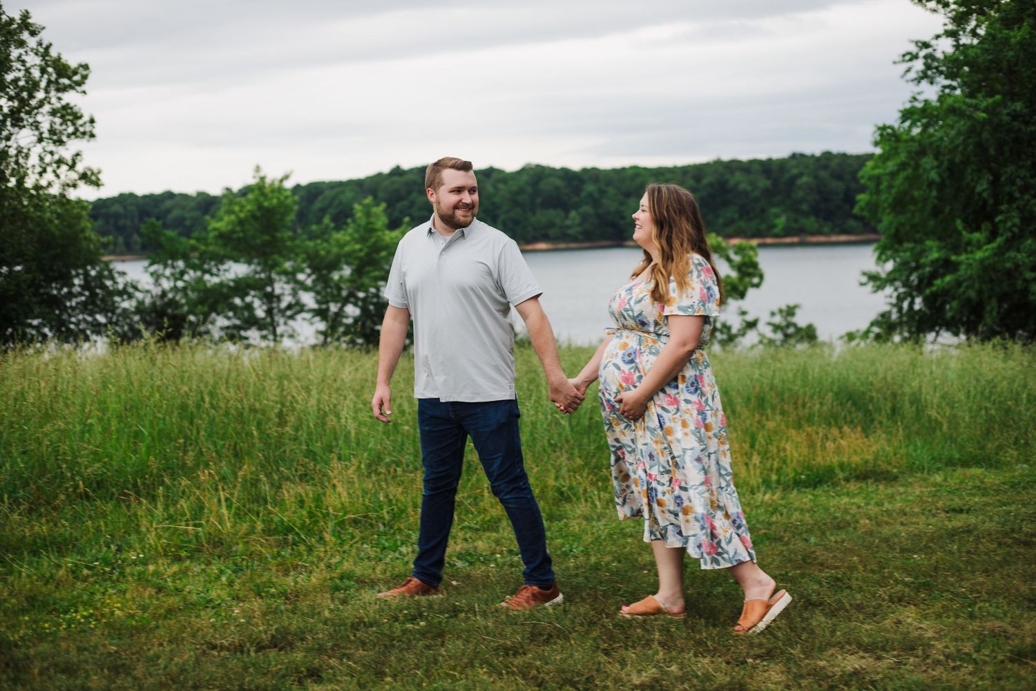Expecting couple holding hands walking in green field near lake during maternity photo session.