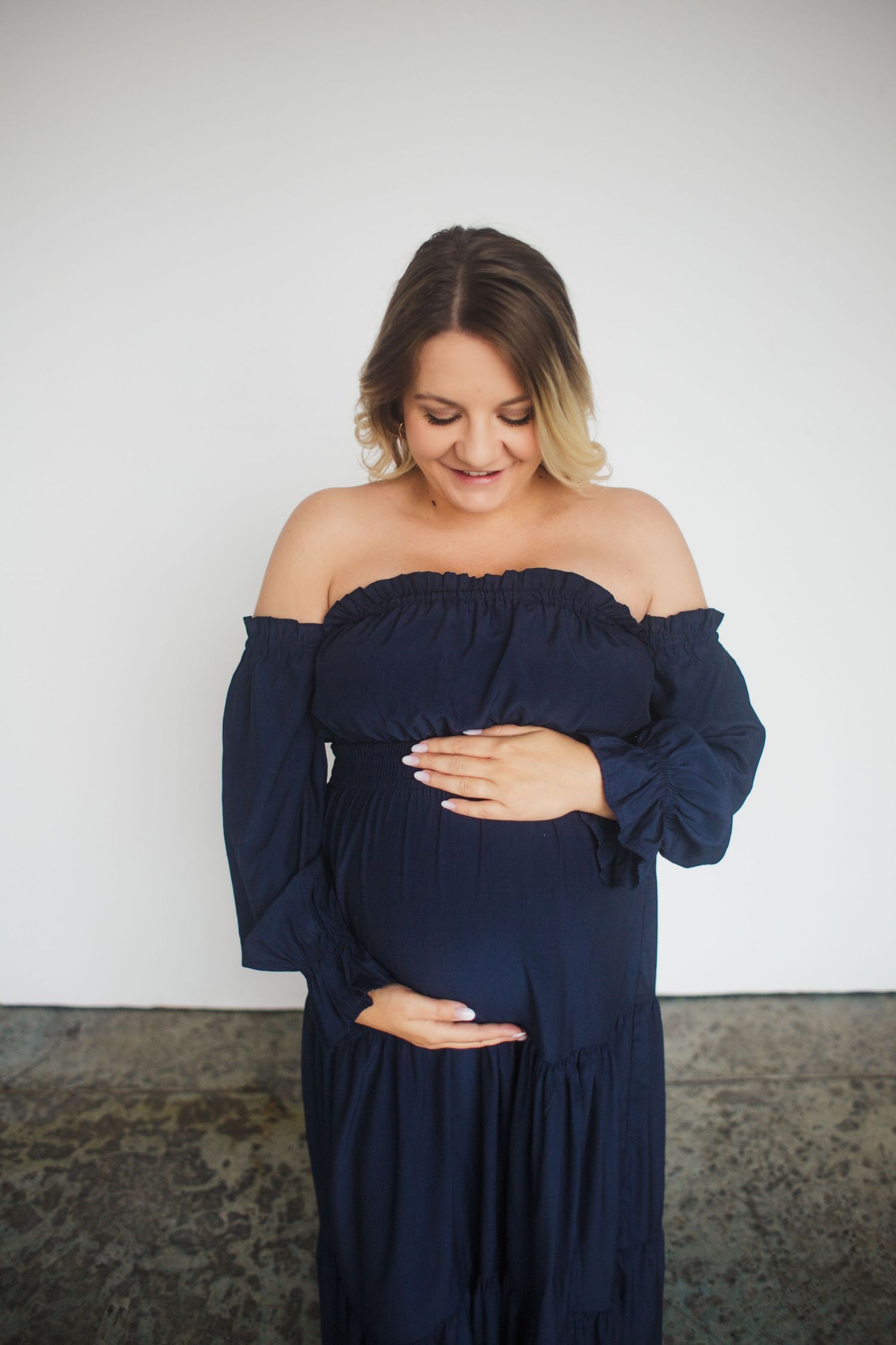 Pregnant woman in navy off-shoulder maxi dress cradling baby bump against white studio wall.