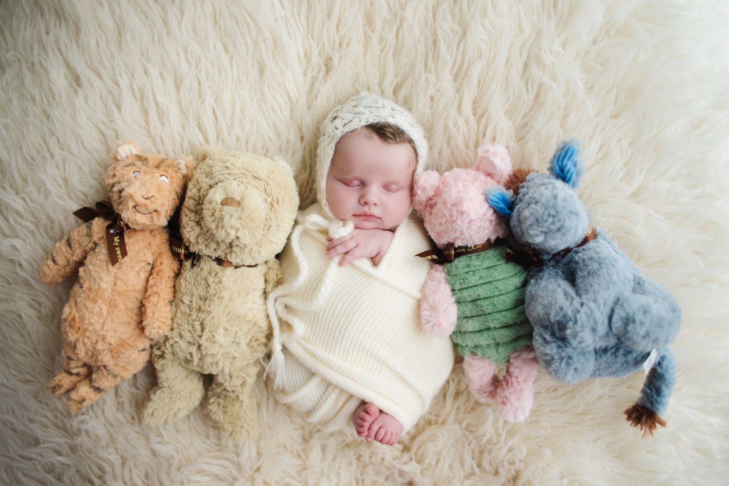 Newborn baby sleeping peacefully between four classic teddy bears on a soft white fur blanket.