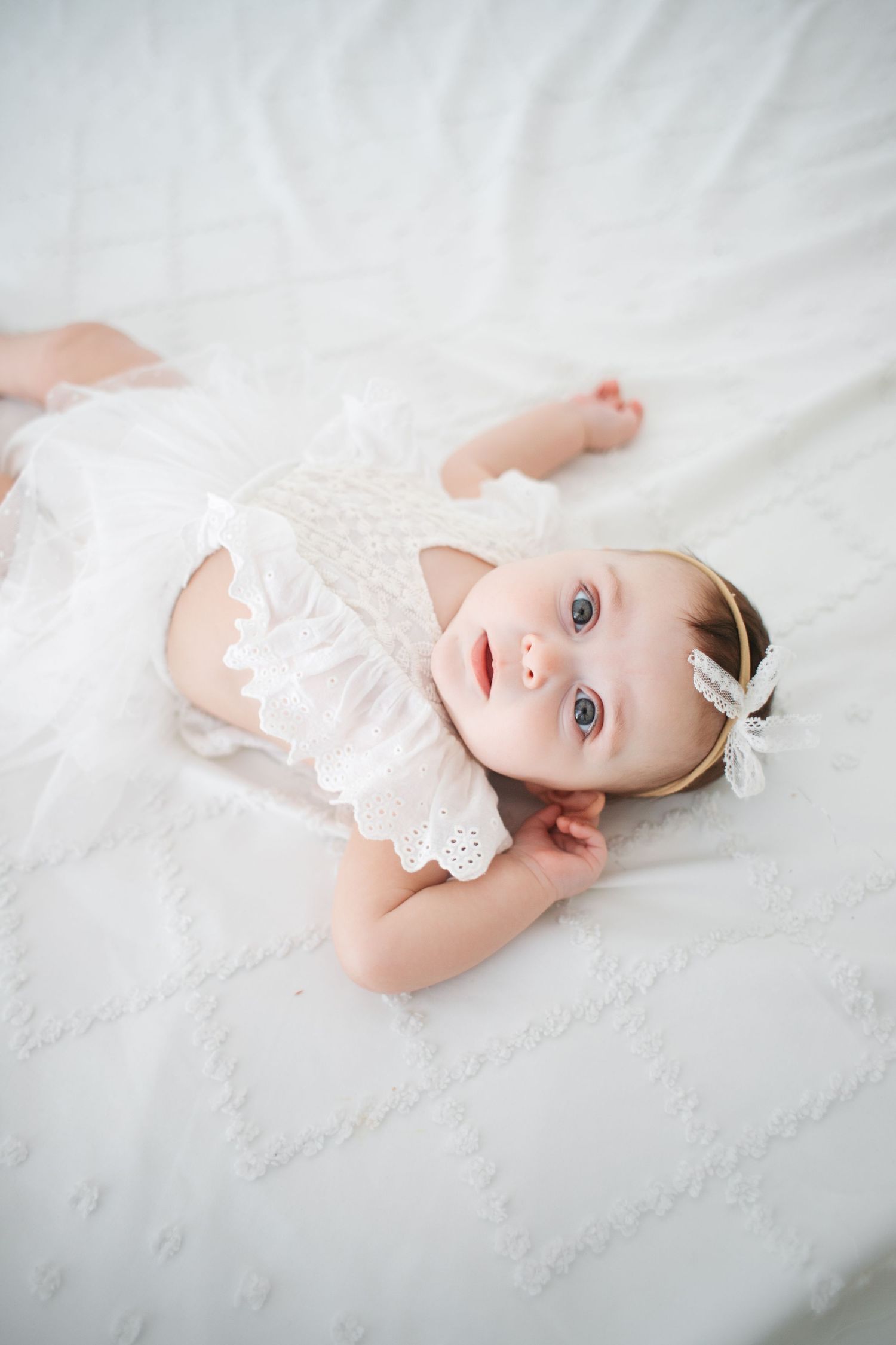 Beautiful baby girl in white lace romper and bow headband lying on white quilted bedding.