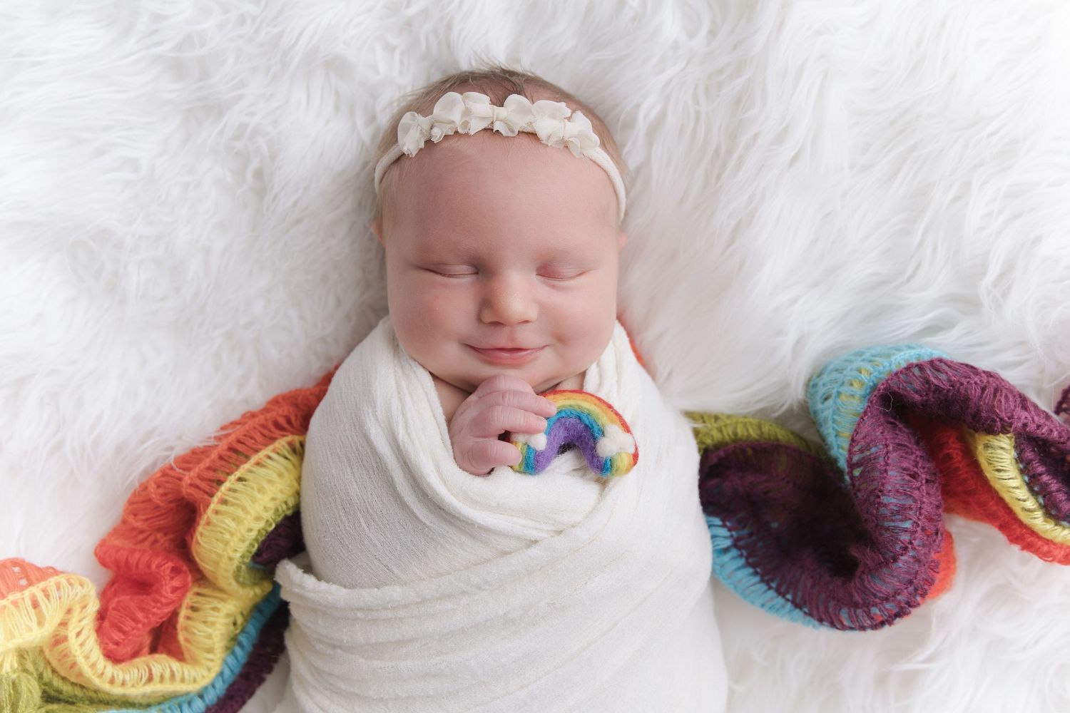 Sleeping newborn baby swaddled in white wrap with rainbow crochet blanket and white floral headband.