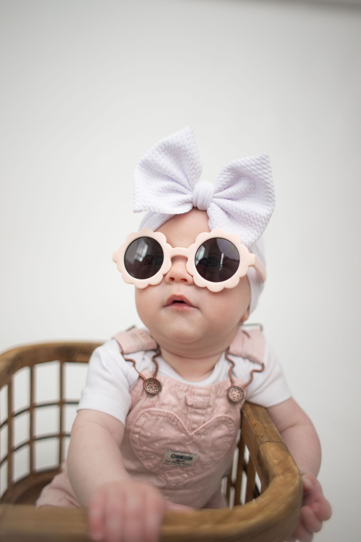 Adorable baby girl wearing pink sunglasses and white bow headband sitting in a wicker chair.