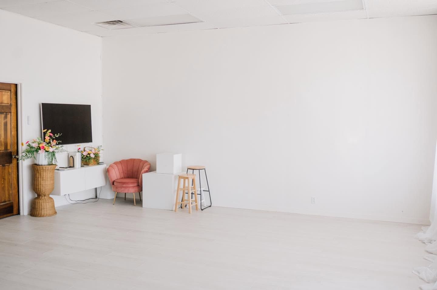 Bright white photography studio with pink velvet chair, wooden stool, TV console, and hardwood floors.