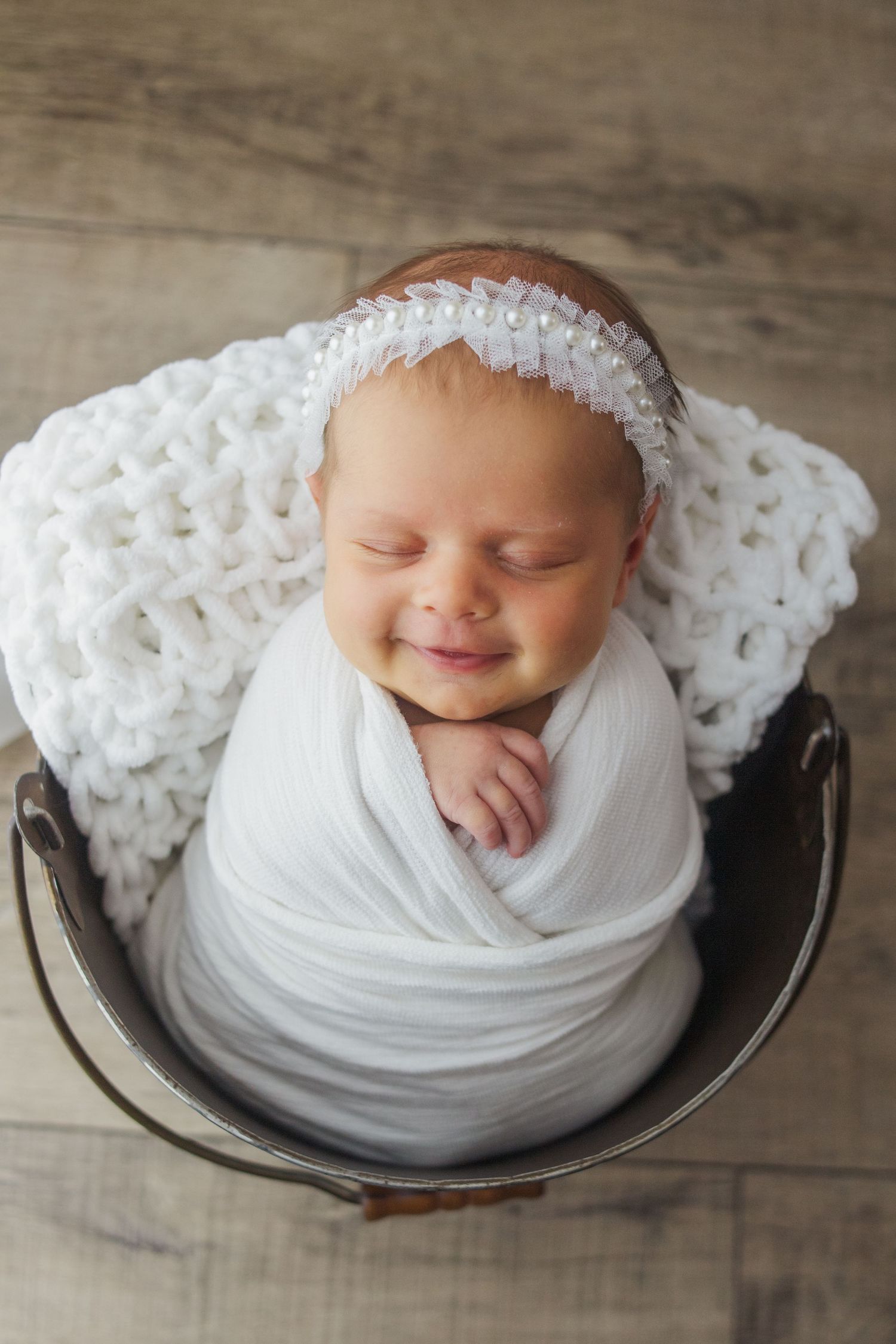 Smiling newborn baby girl swaddled in white wrap sitting in a rustic bucket with pearl headband.