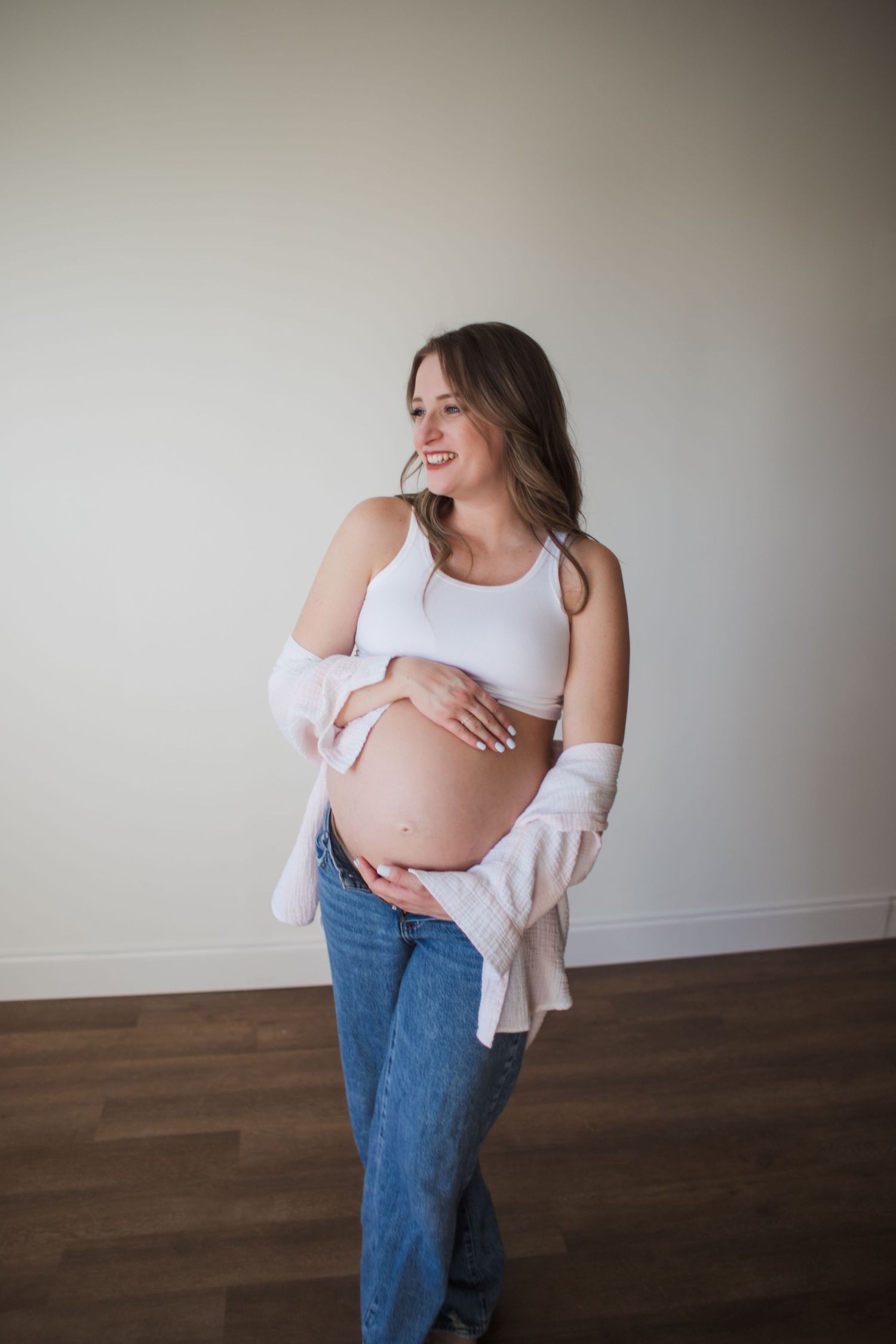 A smiling pregnant woman cradles her baby bump wearing a white top and jeans indoors.