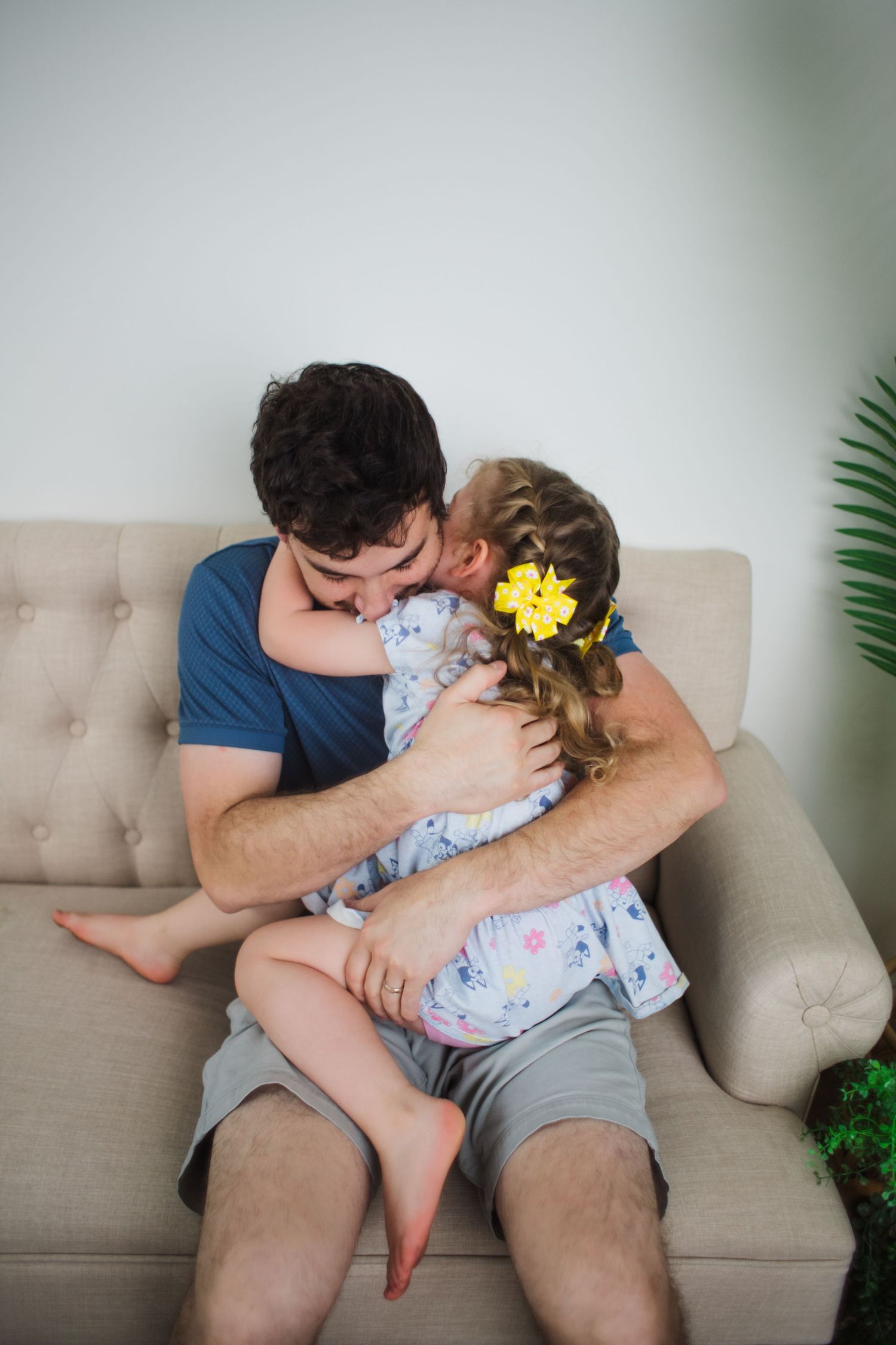 Father tenderly embracing young daughter with yellow bow on beige sofa indoors.