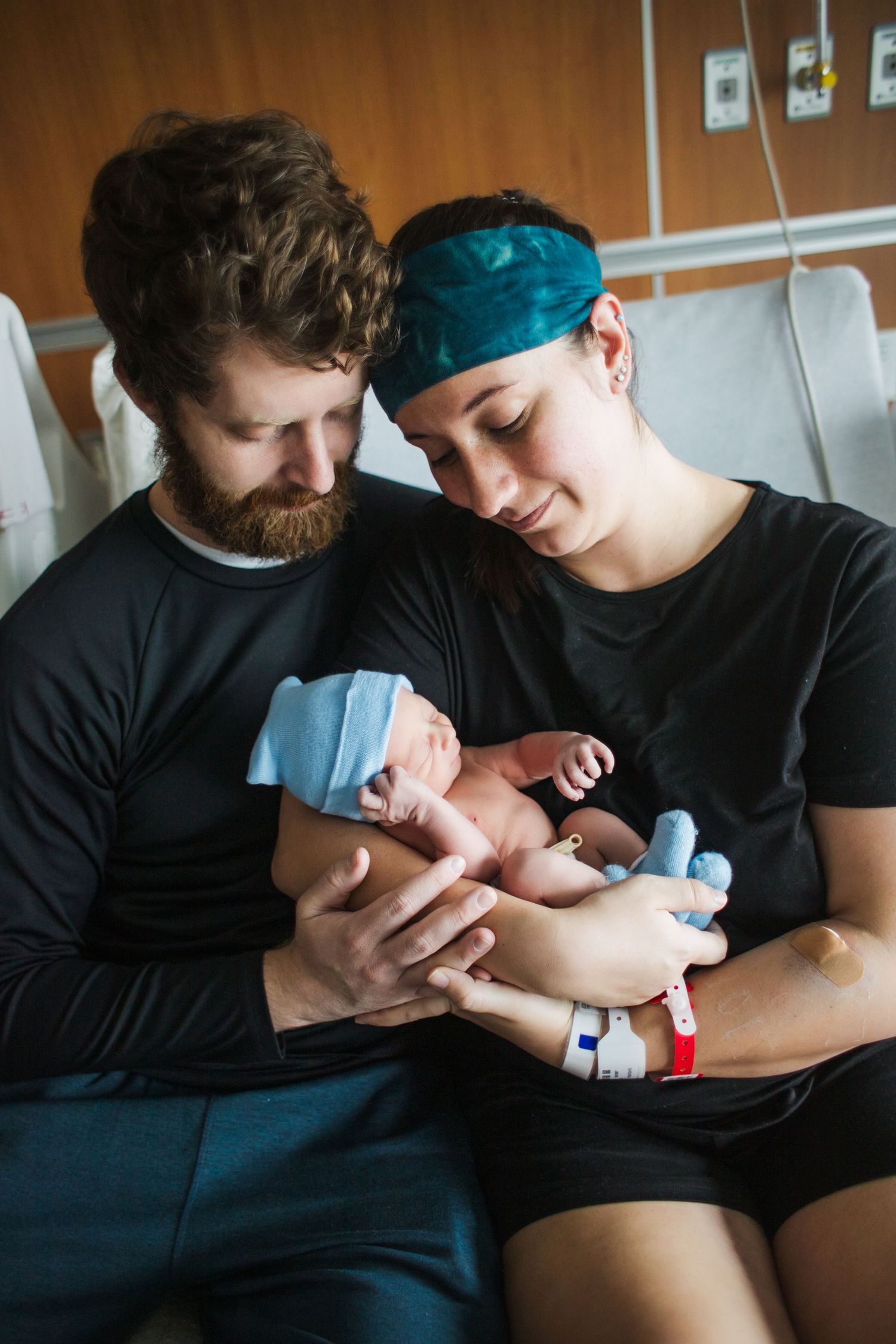 New parents in hospital room lovingly gazing at their swaddled newborn baby together.