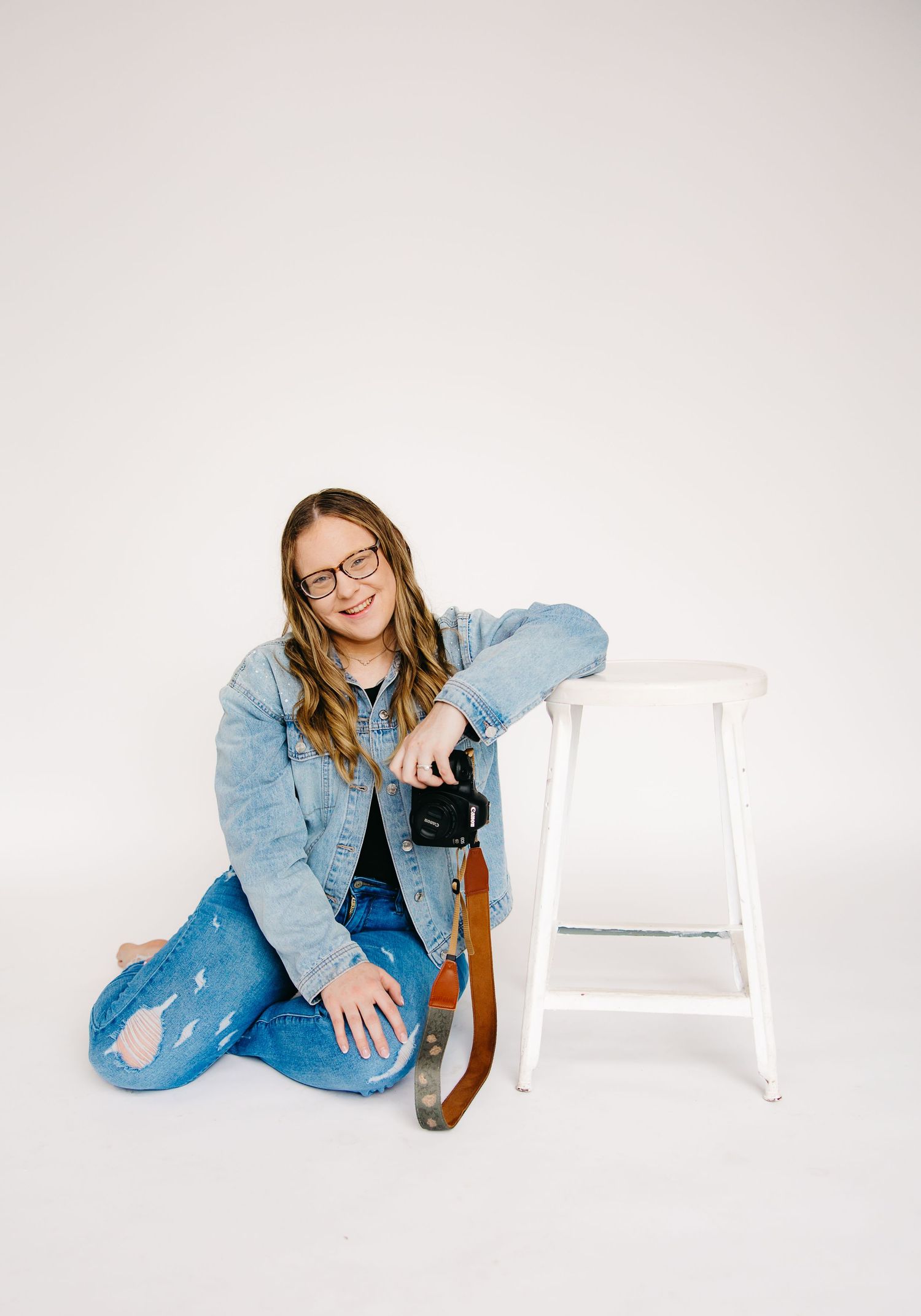 Smiling female photographer sitting on floor beside white stool holding camera, wearing denim jacket and glasses.