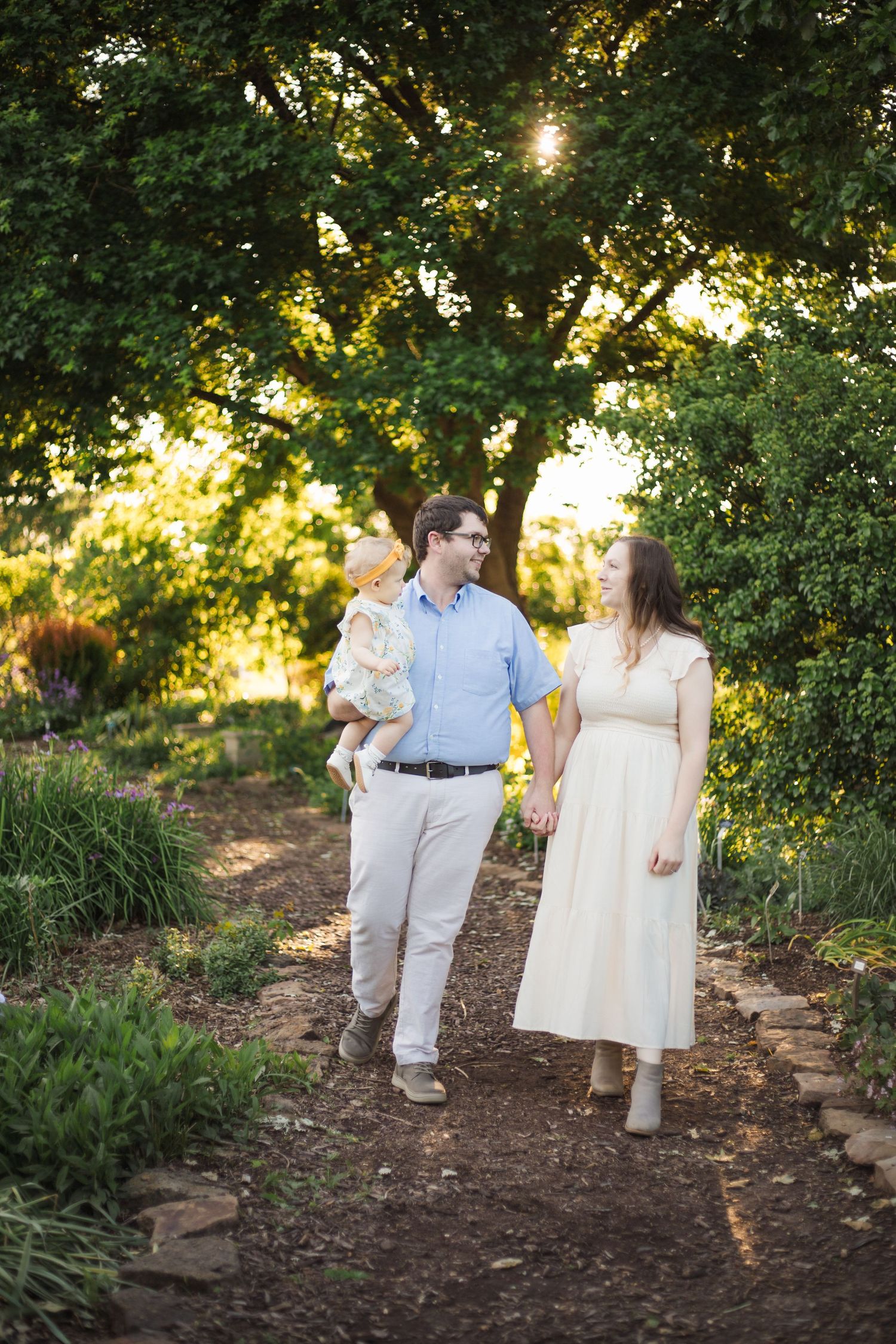A couple walks hand-in-hand carrying their baby through a sunlit garden path.