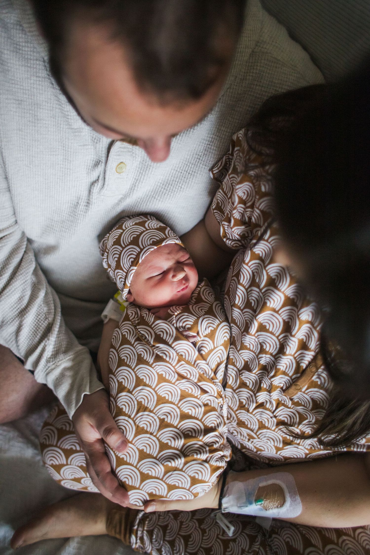 Father holding sleeping newborn dressed in brown patterned outfit and matching hat from above.