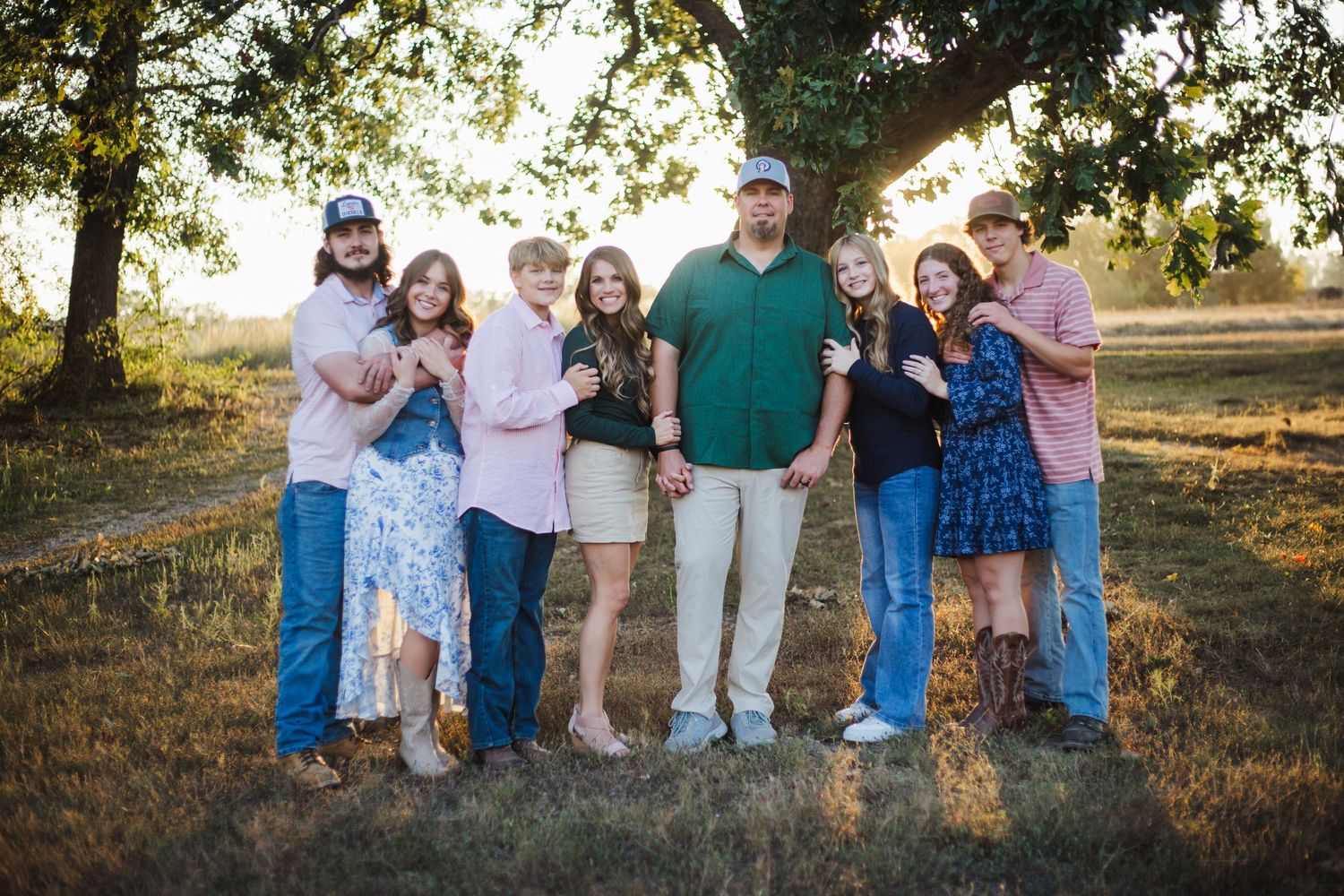 Large extended family of eight adults posing together outdoors under a tree during golden hour sunset session.