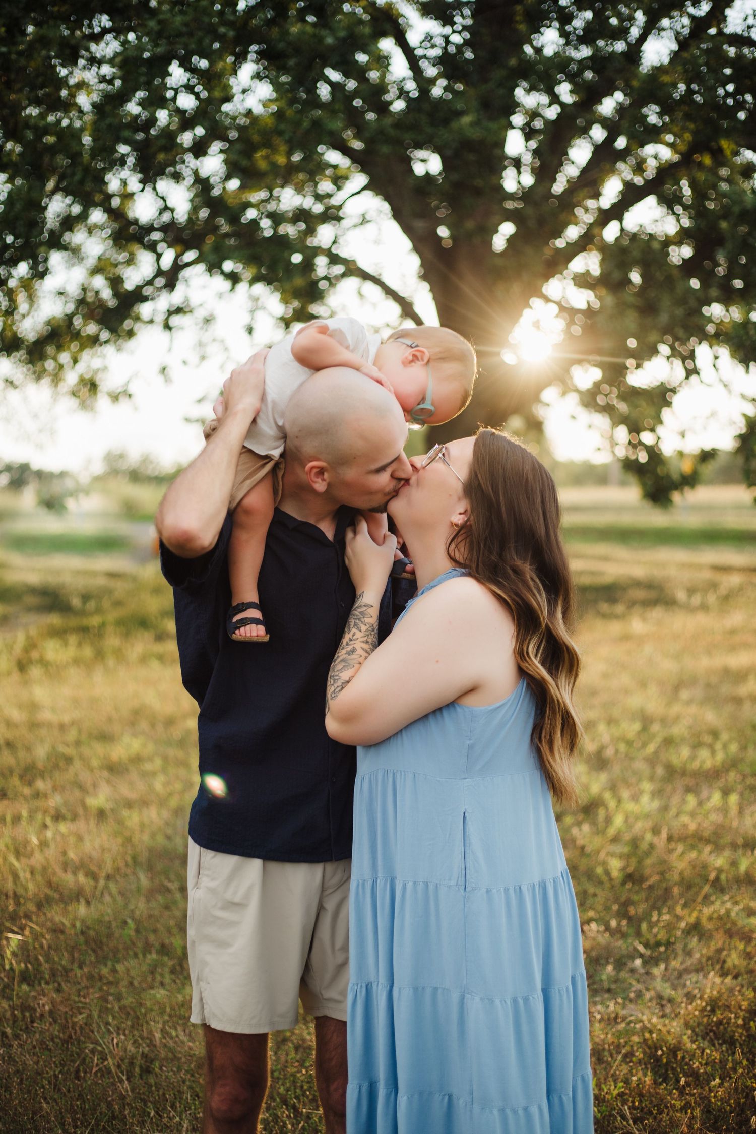 Couple kissing while lifting baby in air during golden sunset in green meadow.