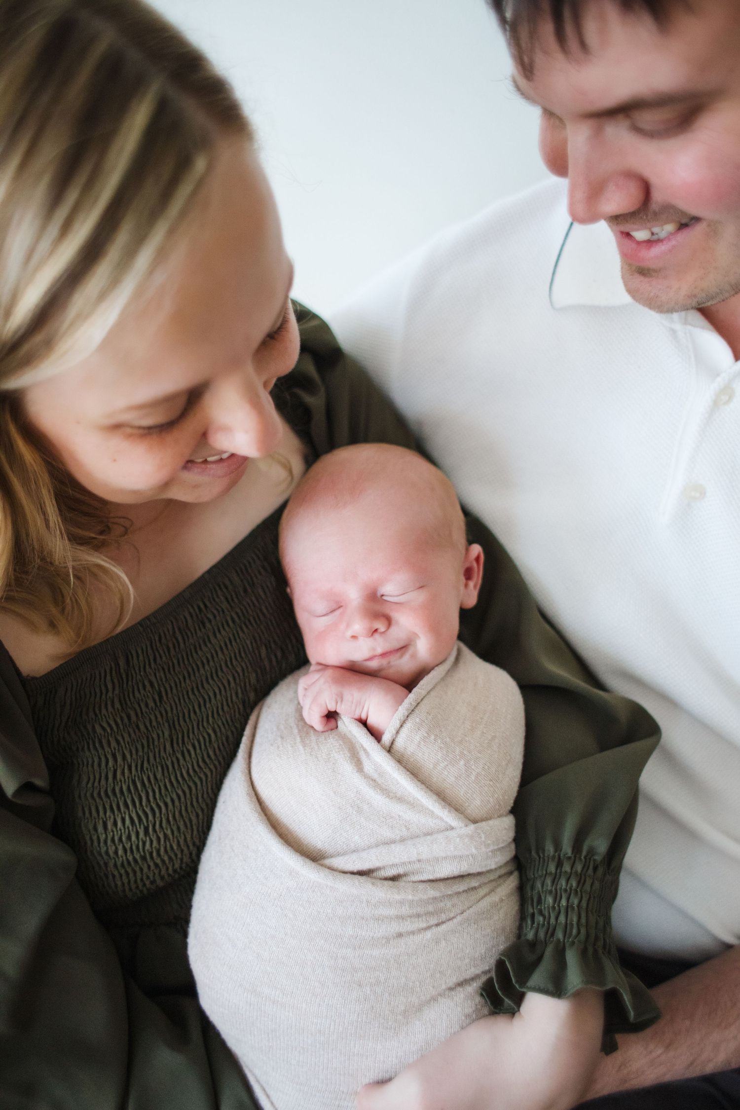 Smiling parents holding their peacefully sleeping swaddled newborn baby wrapped in cream blanket.