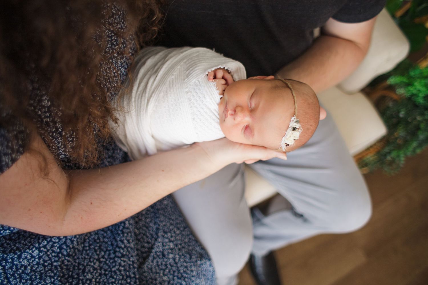 Newborn baby wrapped in white swaddle with floral headband being held by family members.