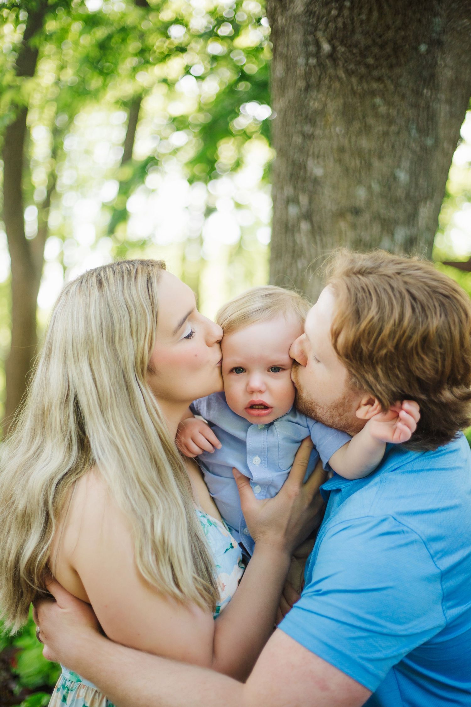 Parents lovingly kiss their baby on the cheeks outdoors in a green park setting.