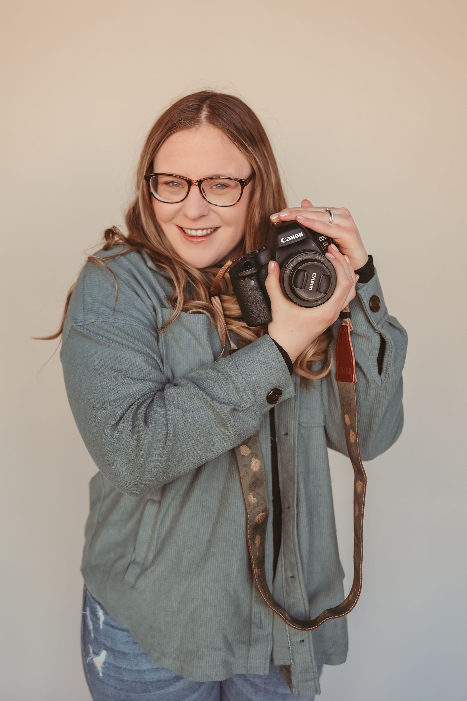 Smiling female photographer holding Canon DSLR camera with leather strap, wearing glasses and gray jacket.