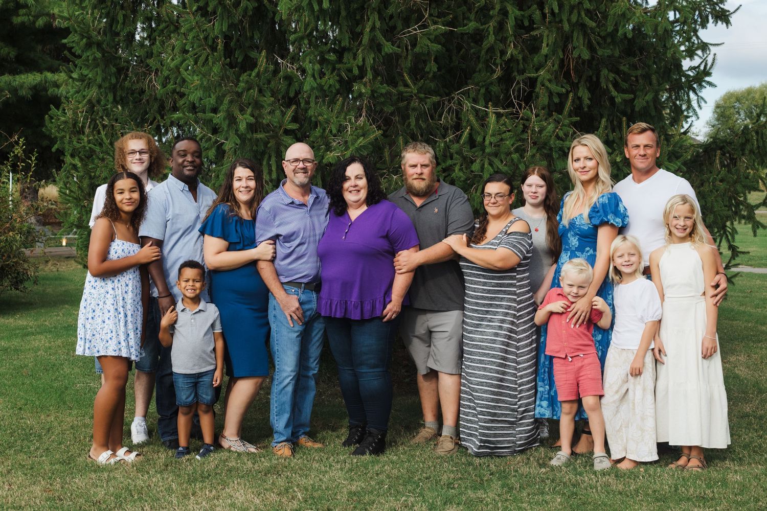 Large multigenerational family group portrait with adults and children smiling together on a green lawn outside.